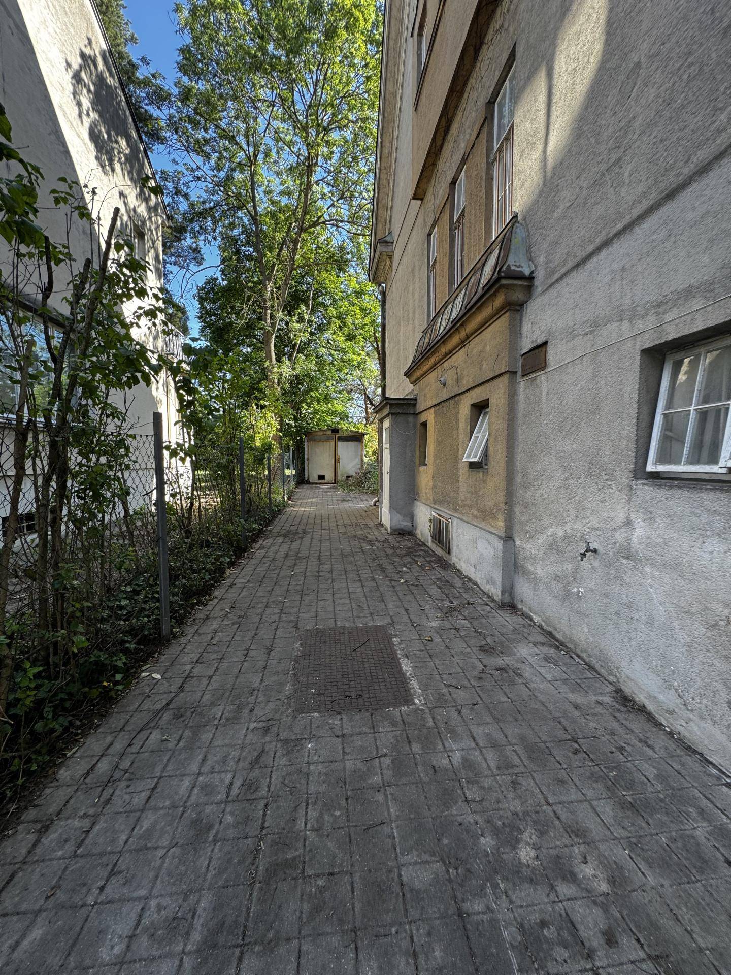 Schmaler gepflasterter Weg entlang der Gebäudeseite mit Bäumen im Hintergrund und viel Grün.