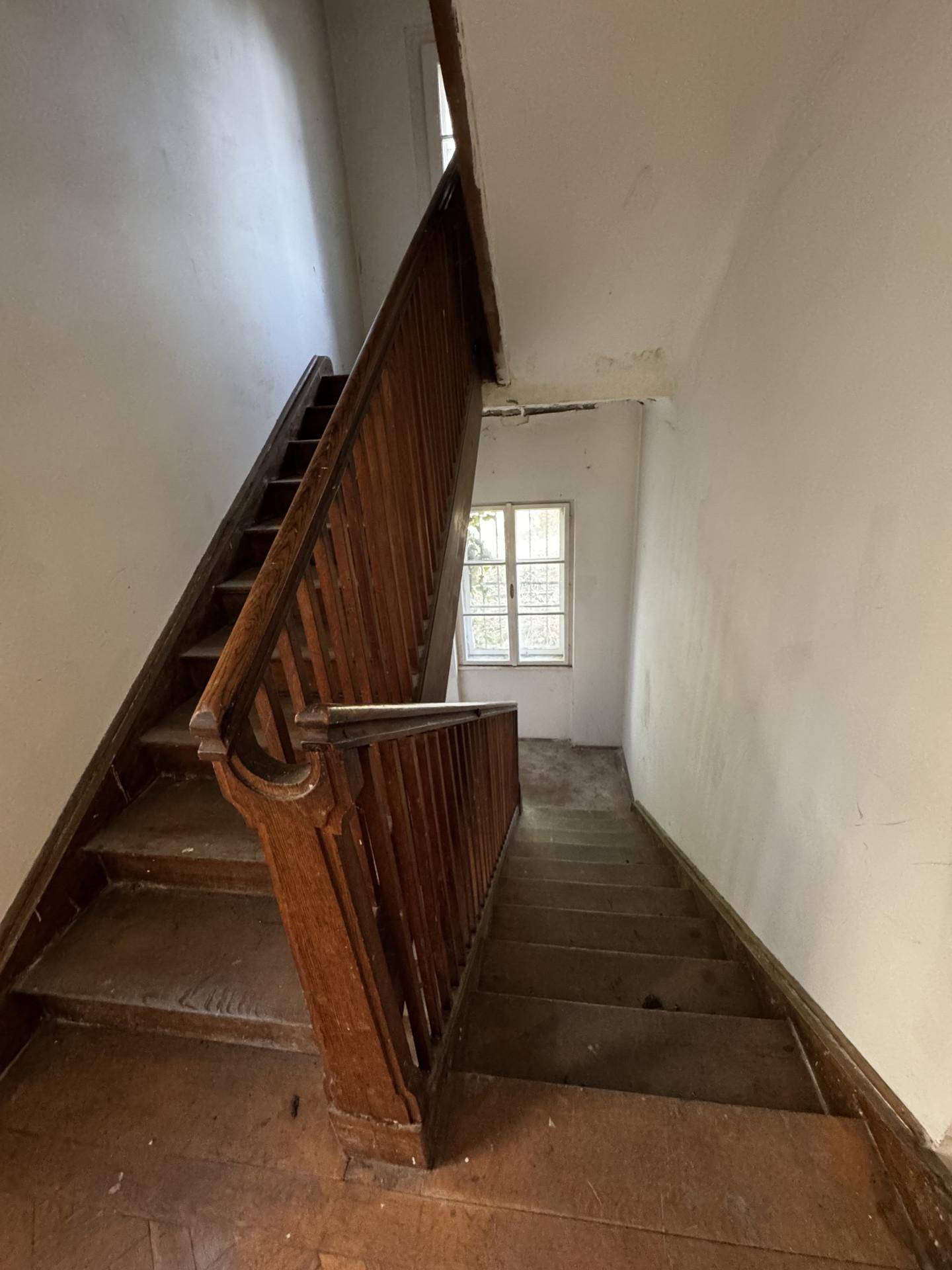 Historische Holztreppe in einem Flur mit Fenster und abgenutztem Holzboden, der saniert werden muss.