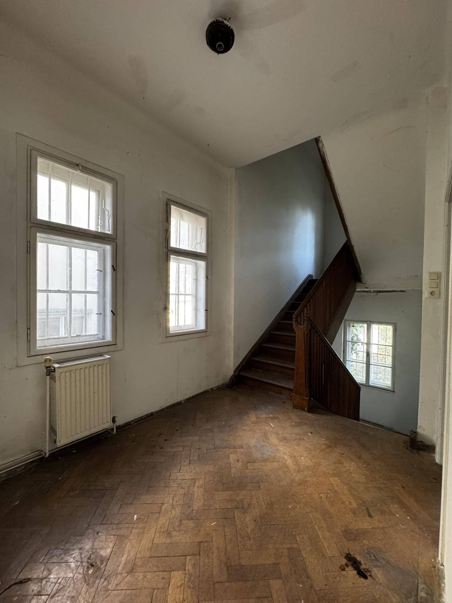 Geräumiger Flur mit Holztreppe, zwei Fenstern und Fischgrätparkett, der renovierungsbedürftig ist.