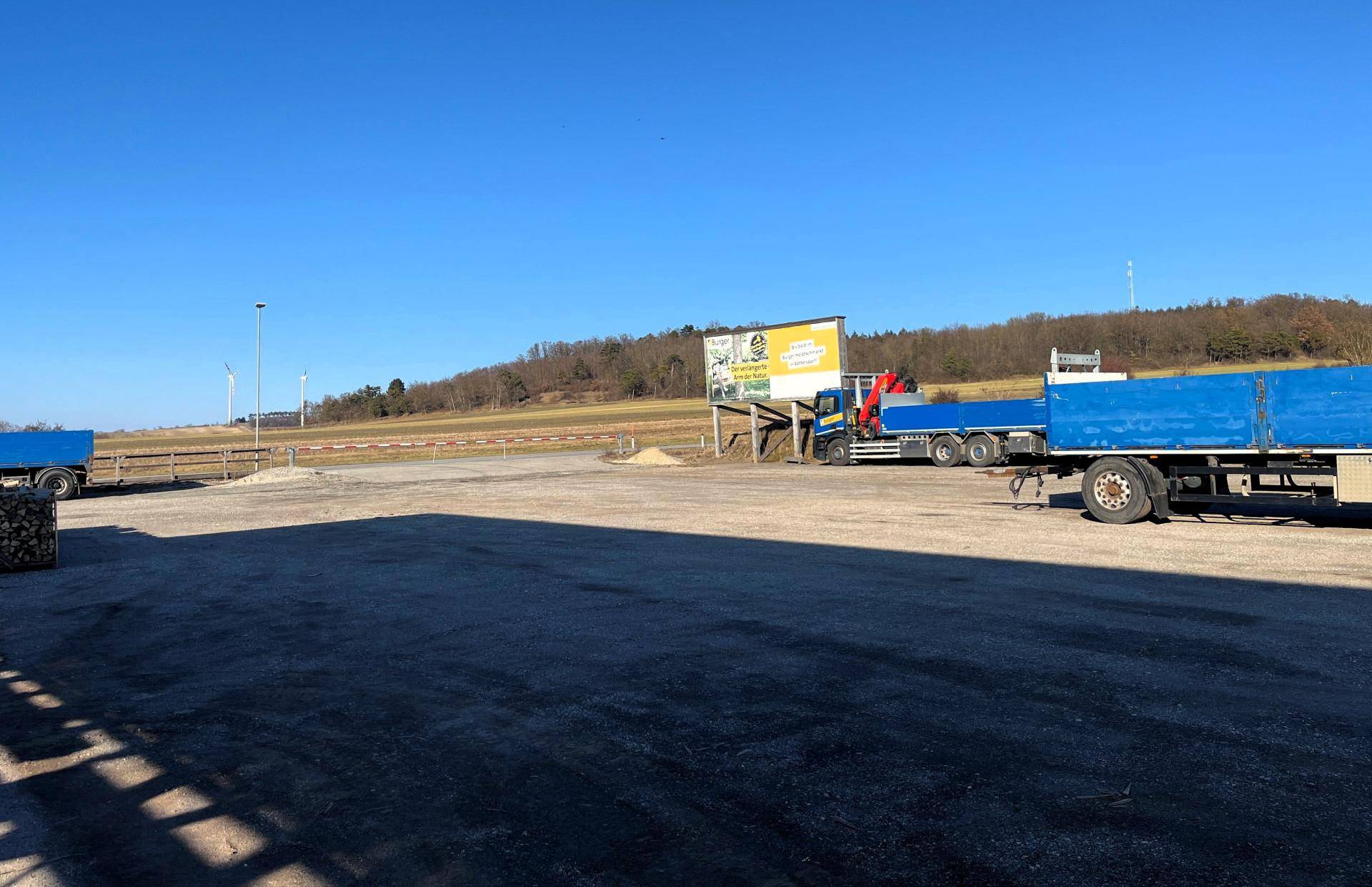 Weitläufiger Schotterplatz mit parkenden Lastwagen und einem Werbeschild, umgeben von Natur und Hügeln.