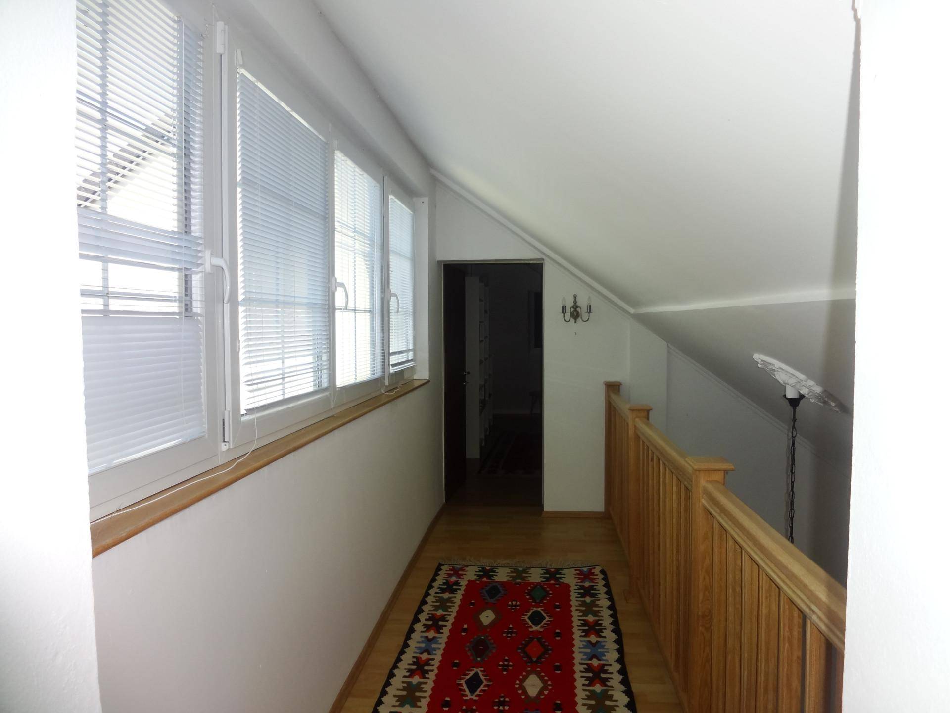 Heller Flur im Obergeschoss mit Holzgeländer, vielen Fenstern und Teppichboden.