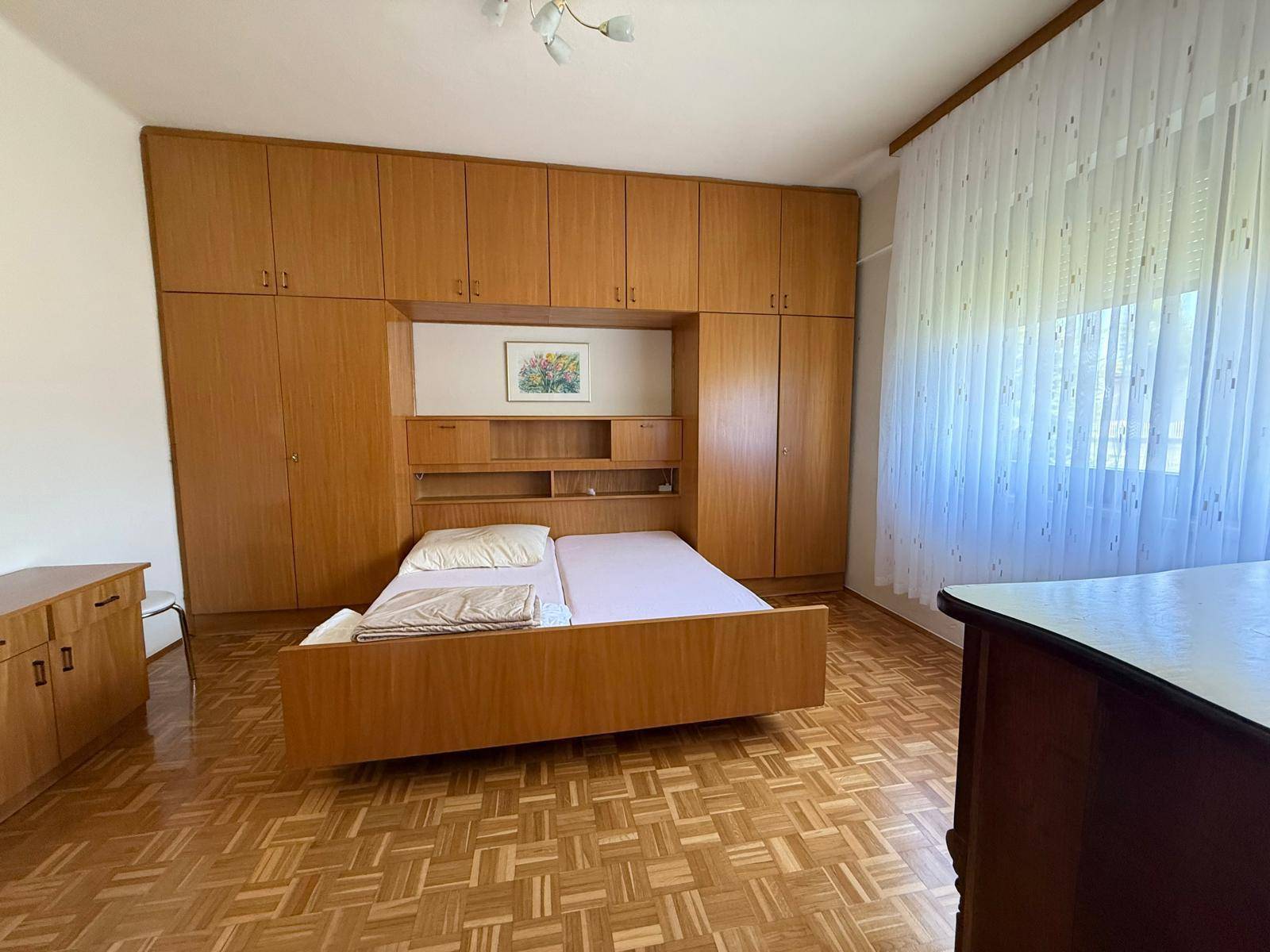 Schlafzimmer mit großem Holzeinbauschrank, Doppelbett und Parkettboden, hell durch das Fenster.