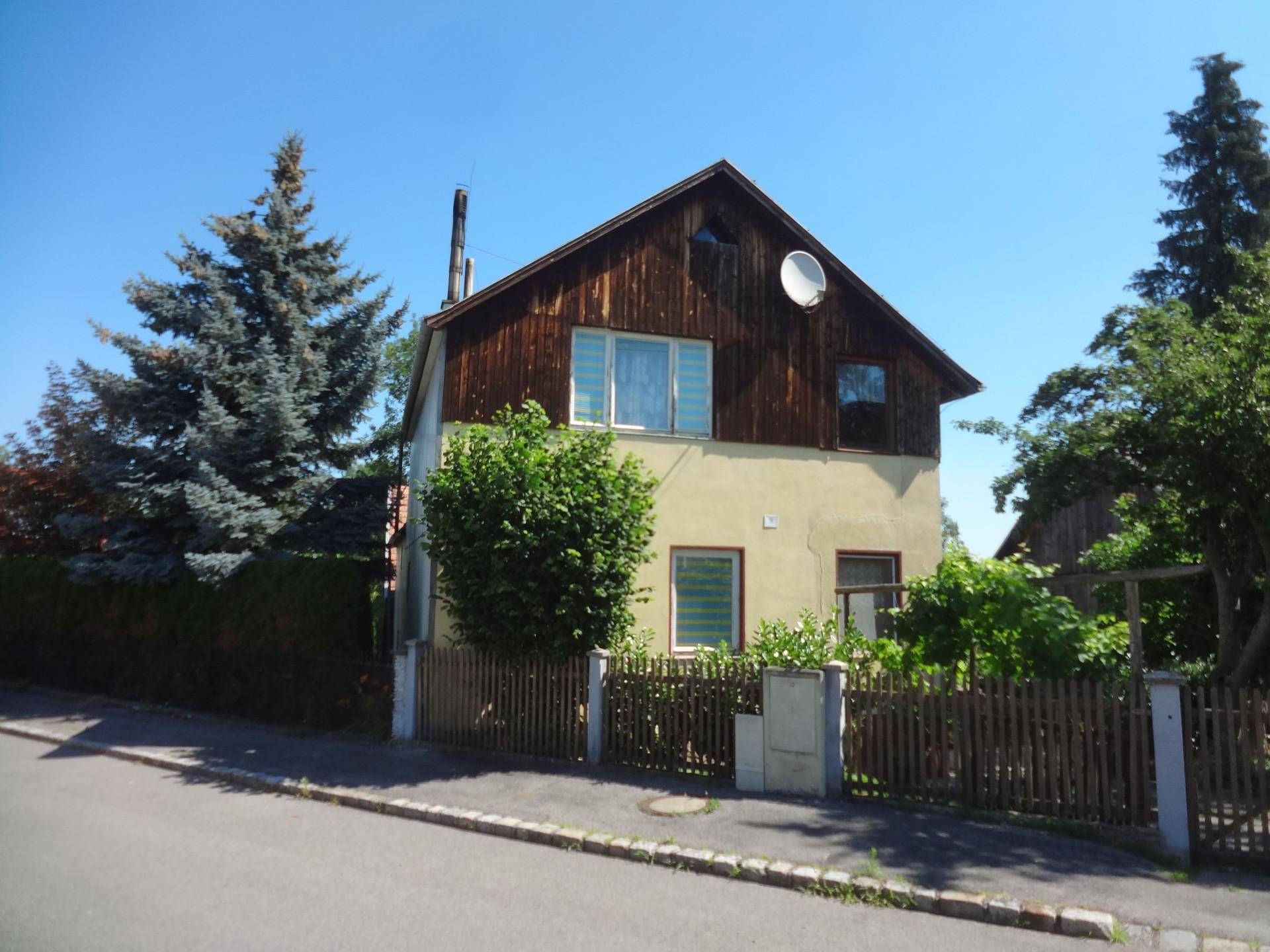 Vorderansicht des Hauses mit Holzdach, Zaun und großen Nadelbäumen.