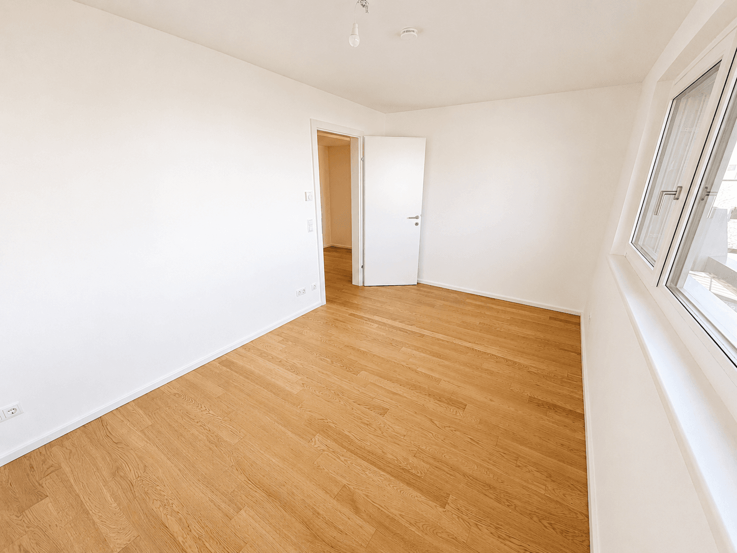 Unmöbliertes Zimmer mit Holzboden, Fenster und Zugang zu einem weiteren Raum.