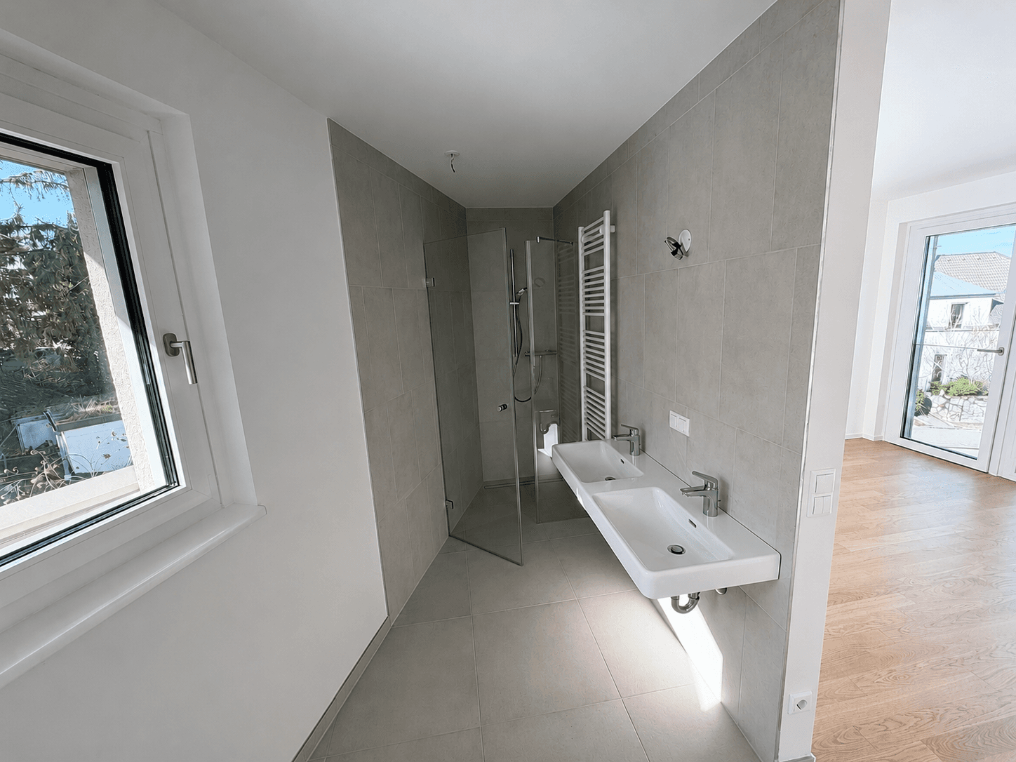 Modernes Badezimmer mit grauen Fliesen, begehbarer Dusche, Doppelwaschbecken und Fenster.