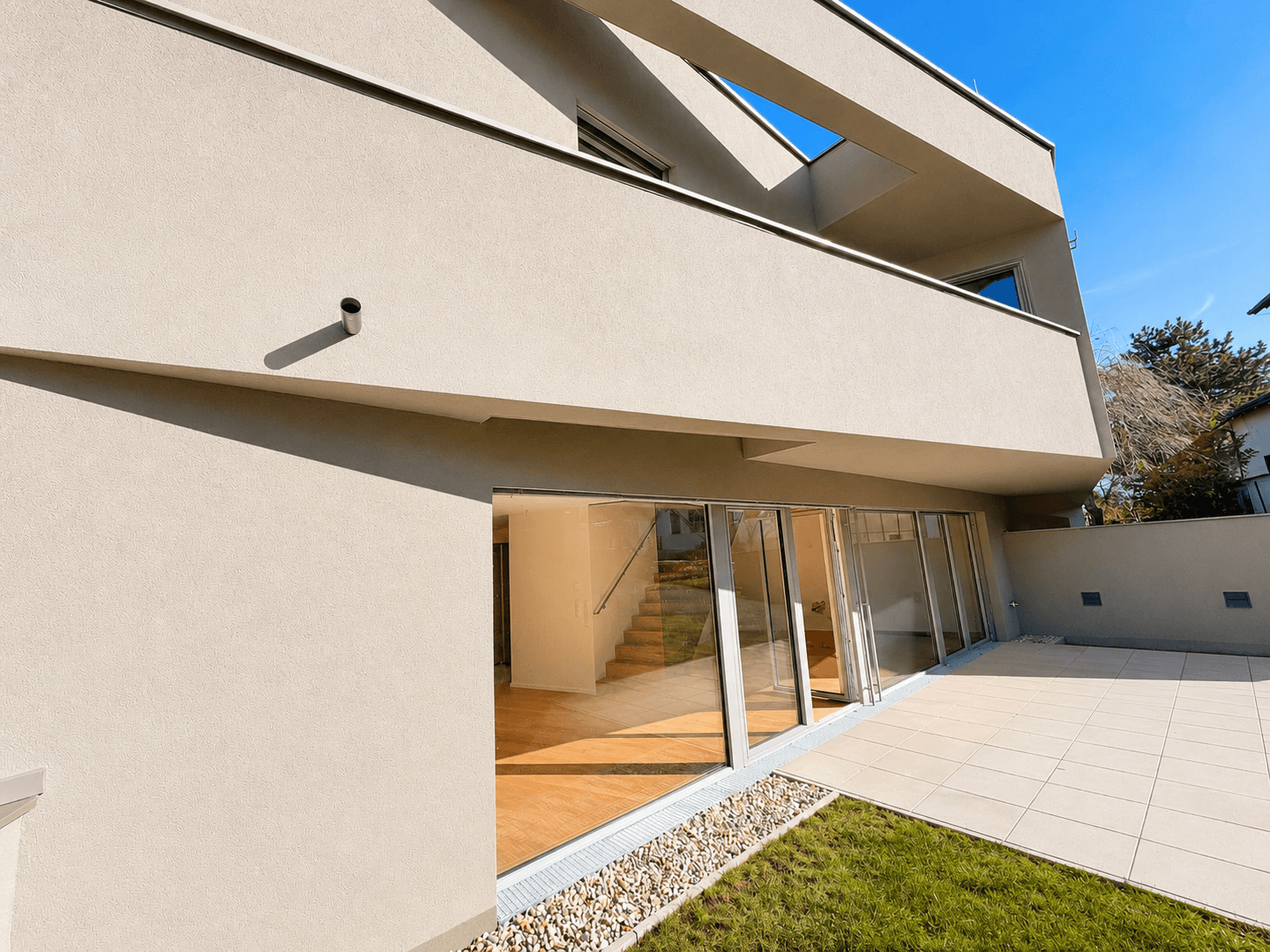 Moderne Architektur mit großer, gefliester Terrasse und angrenzendem Rasenbereich unter blauem Himmel.