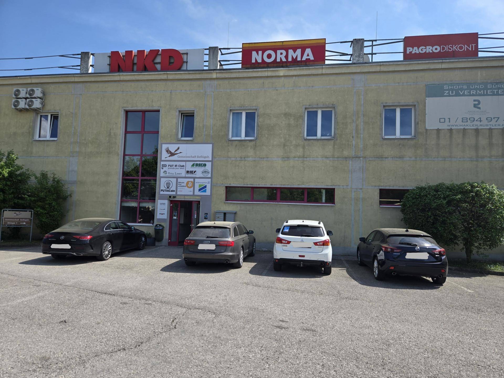 Fassade eines Geschäftshauses mit mehreren Einzelhandelsgeschäften und Parkplätzen davor.