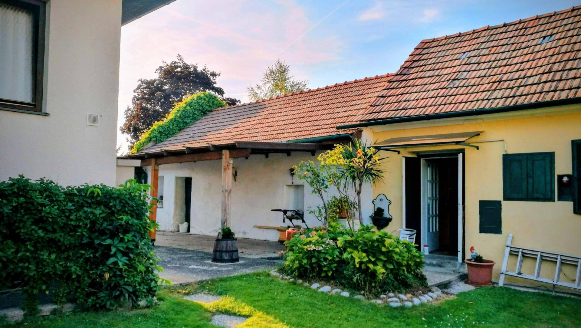 Rustikales Gebäude mit Ziegeldach, überdachtem Terrassenbereich und einem kleinen, bepflanzten Garten.