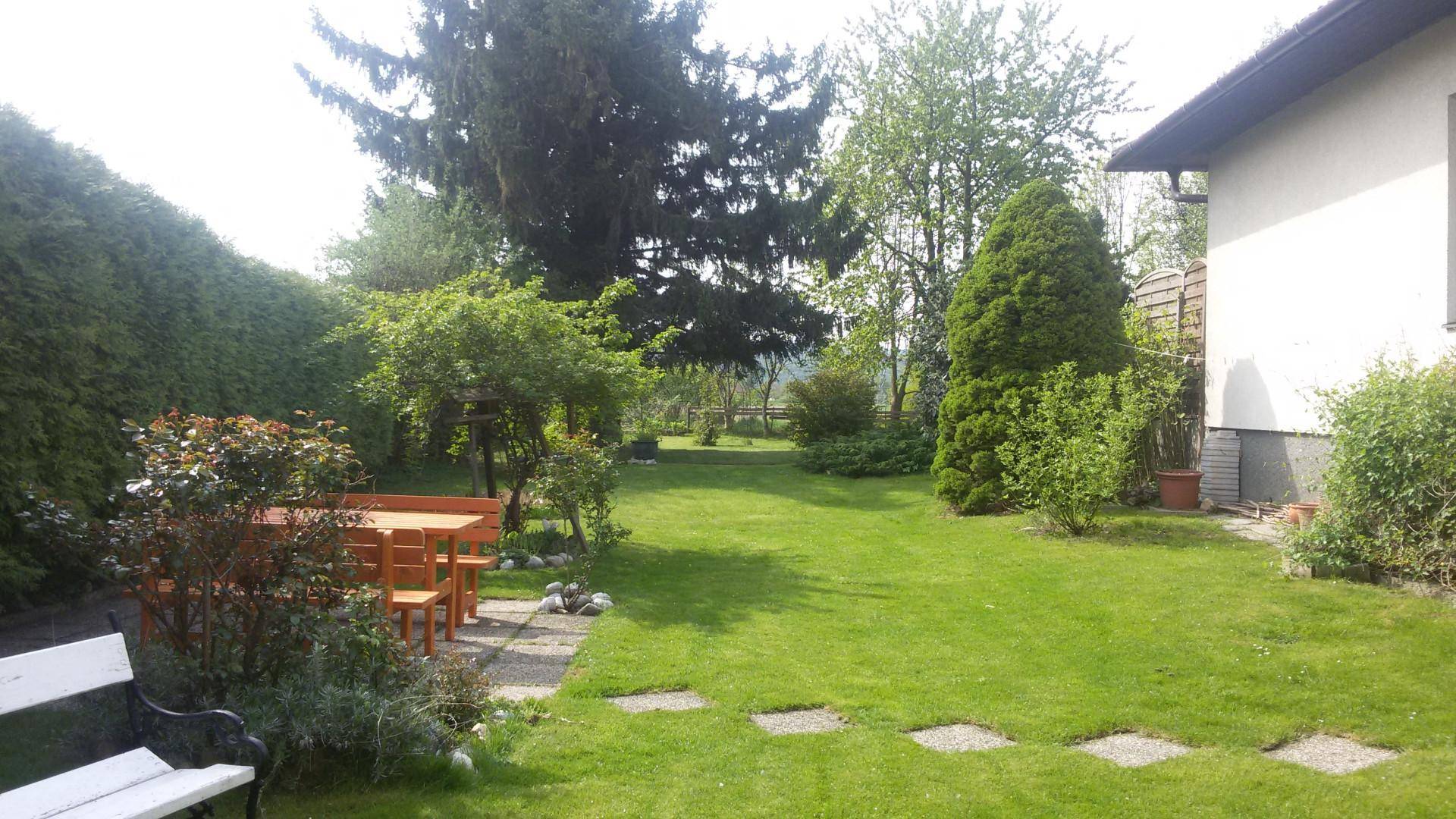 Großer grüner Garten mit Holztisch und Bänken, umgeben von Bäumen und Hecken.