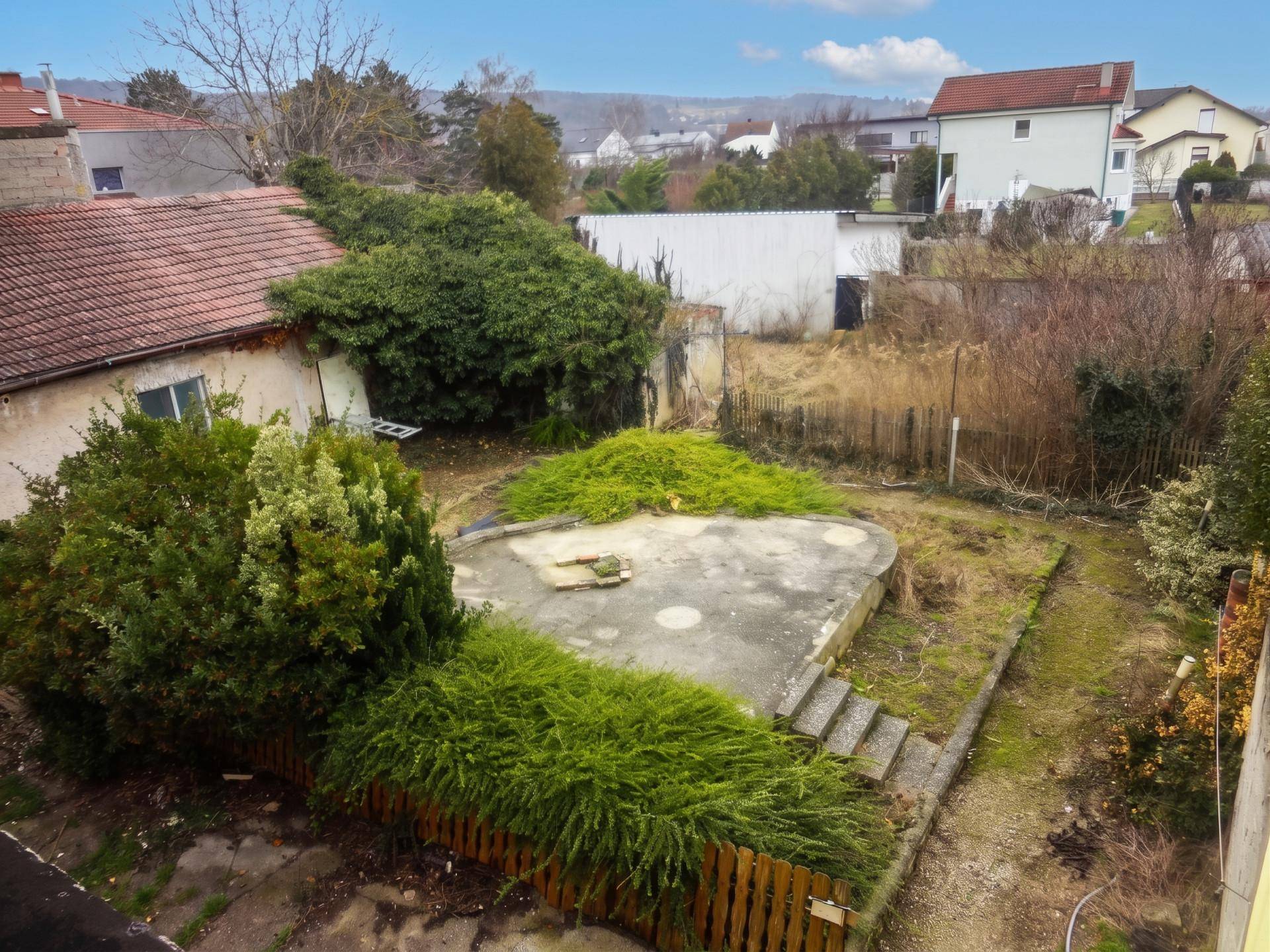 Blick von oben auf einen verwilderten Garten mit einer runden Betonstruktur und viel Grün.