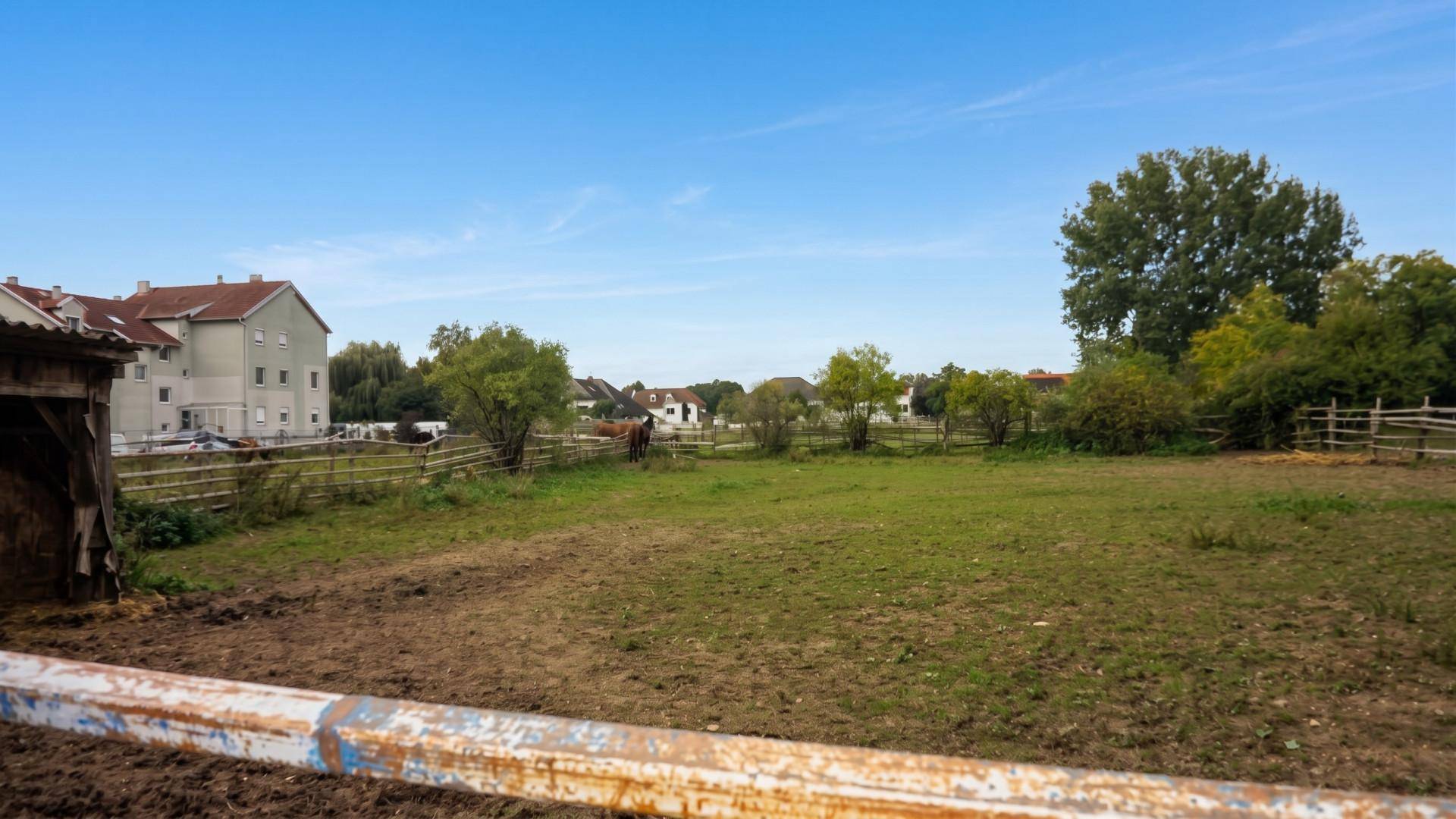 Offenes Feld mit Zaun im Vordergrund und Wohngebäuden in der Ferne unter klarem Himmel.