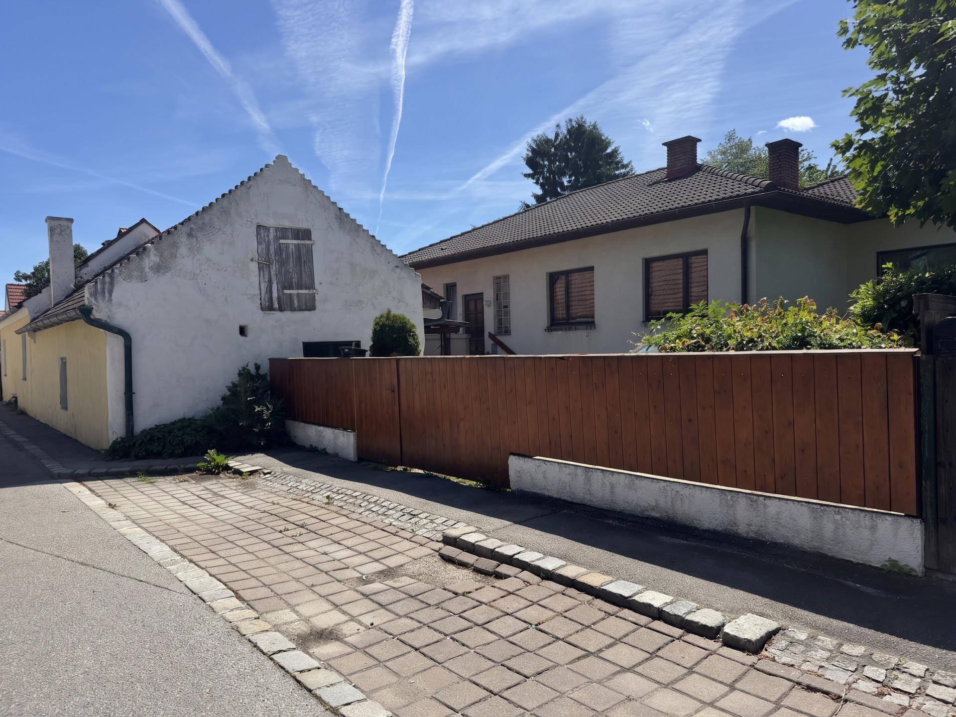 Straßenansicht mit zwei Häusern, einem älteren und einem neueren, und einem Holzzaun im Vordergrund.