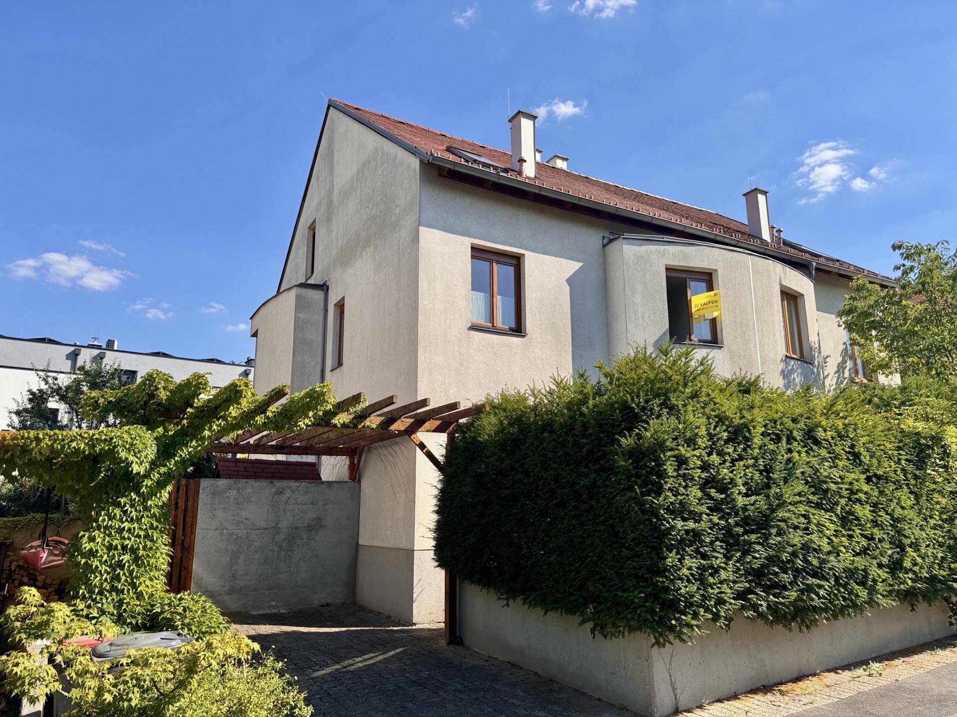 Hausfassade mit Pergola und Hecke unter blauem Himmel an einem sonnigen Tag.