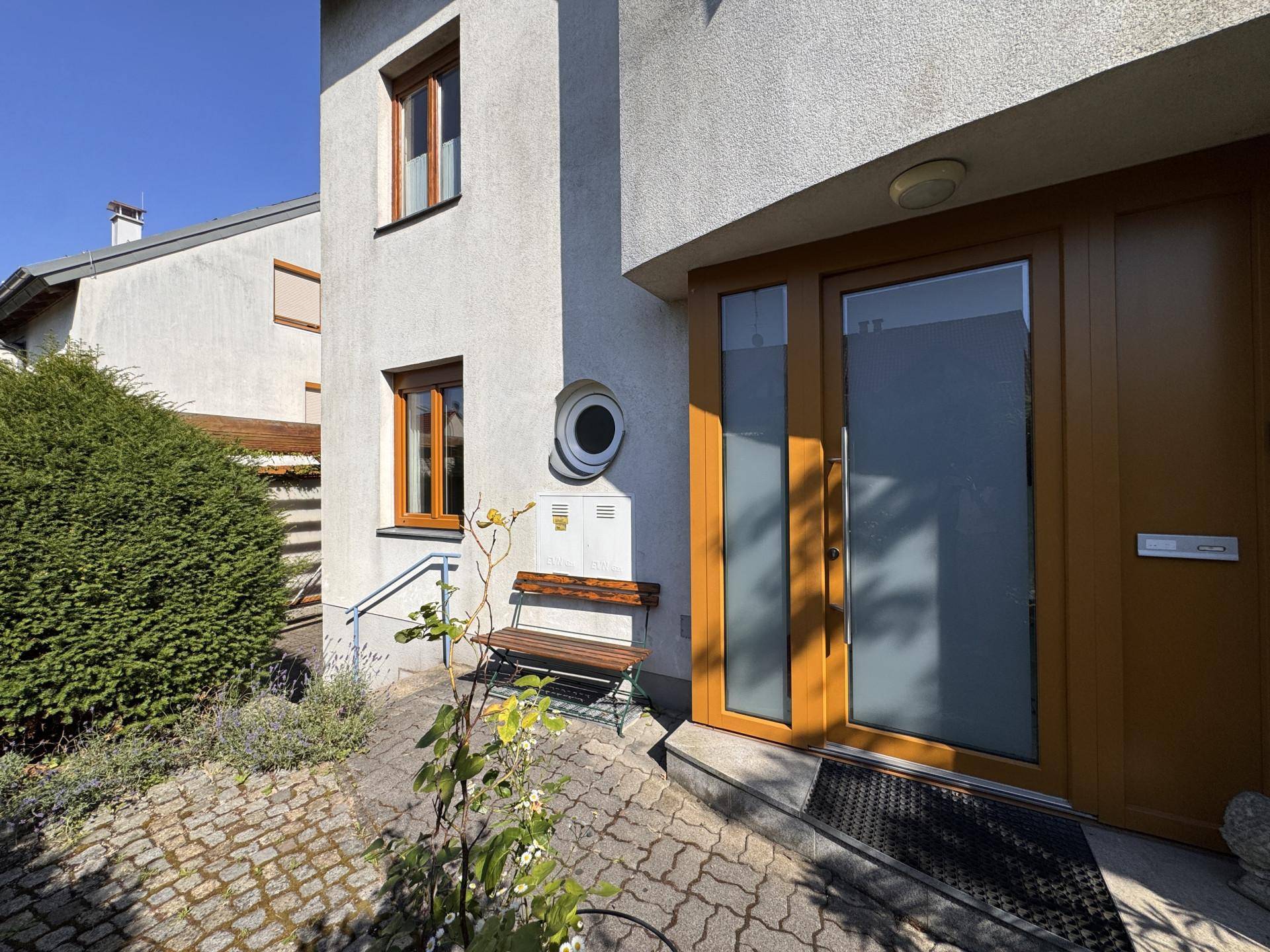 Hauseingang mit Holztür, Seitenfenstern und kleiner Sitzbank im Vorgartenbereich.