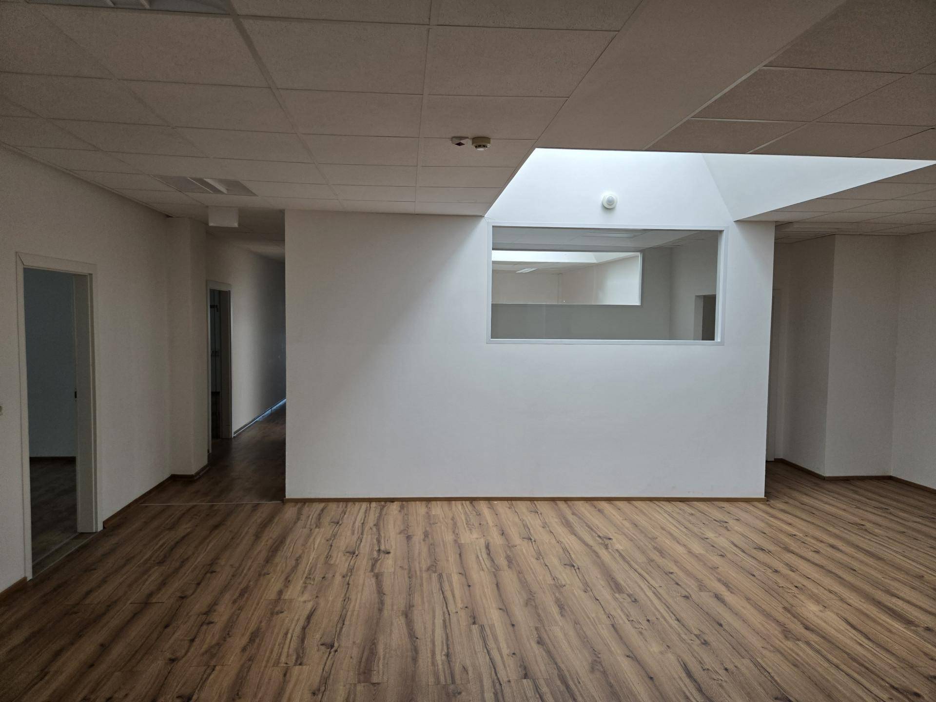 Großer Büroraum mit Laminatboden, abgehängter Decke und einer internen Fensteröffnung.