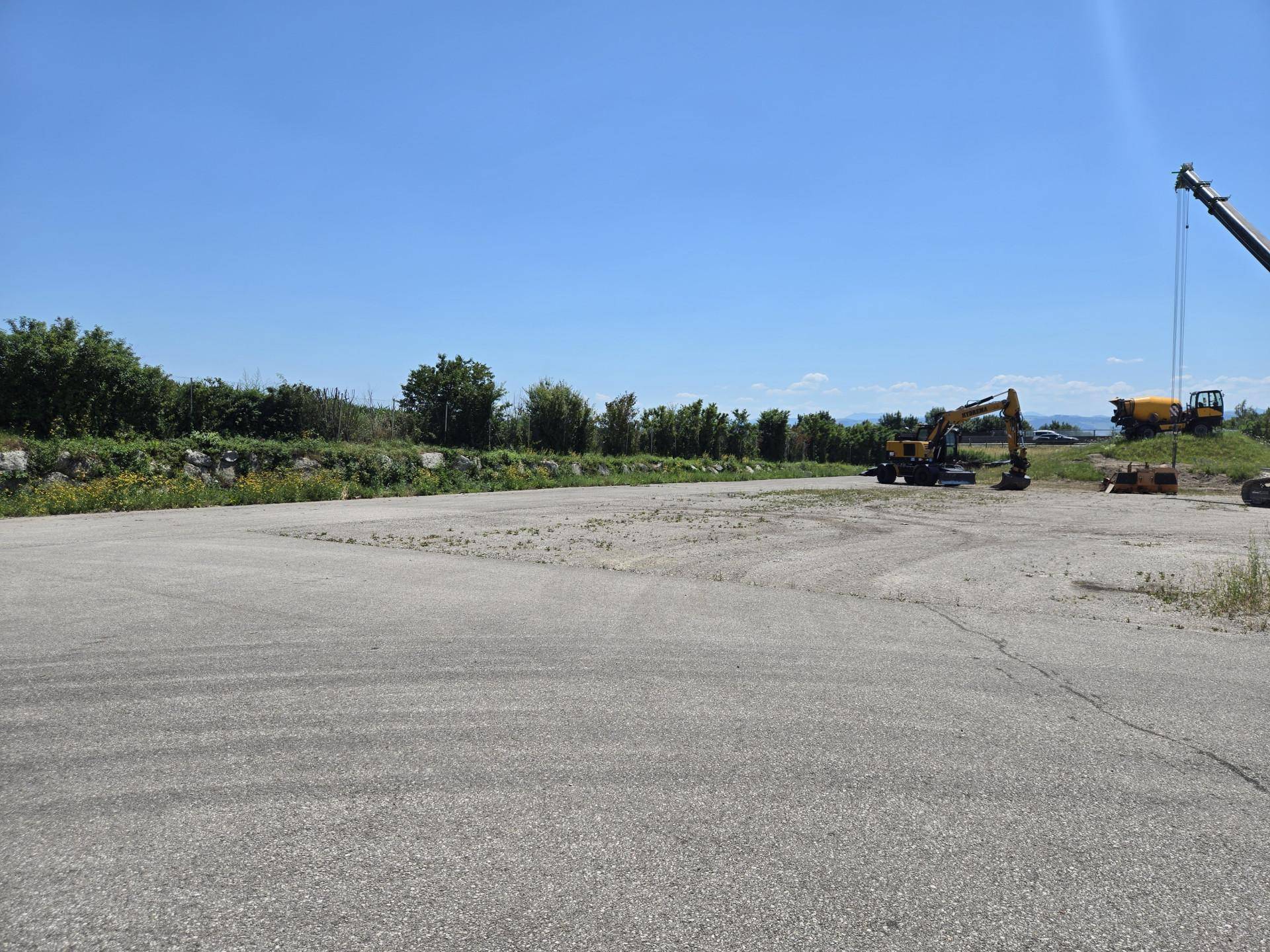 Weitläufige, asphaltierte Fläche mit Bagger und Kran im Hintergrund, umgeben von Vegetation und Bergen.