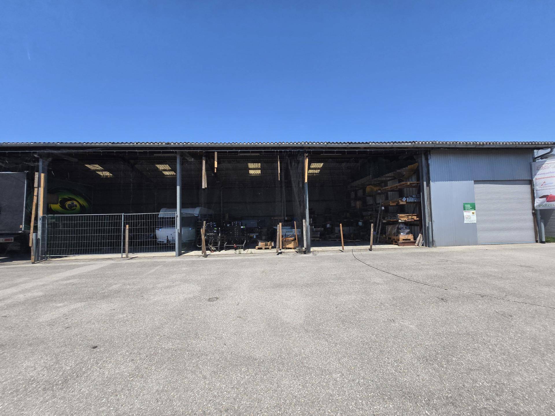 Offene Lagerhalle mit verschiedenen Materialien und Geräten auf einem gepflasterten Vorplatz bei Tageslicht.