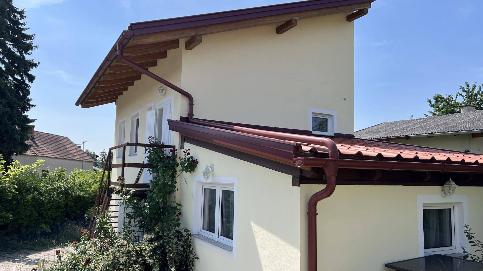 Seitenansicht des Hauses mit externer Holztreppe und gelber Fassade unter blauem Himmel.