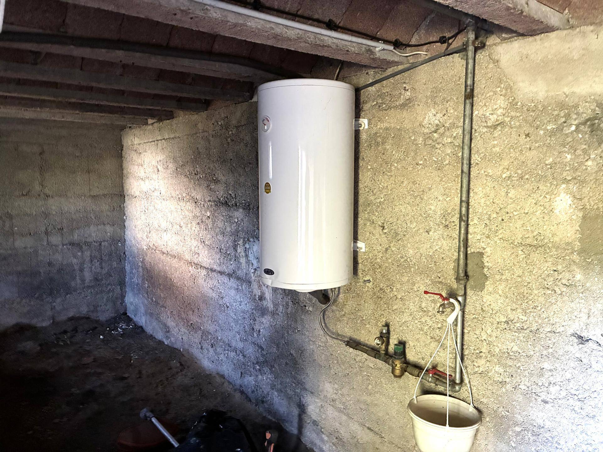 Kellerraum mit Warmwasserboiler, sichtbaren Leitungen und unverputzten Wänden.