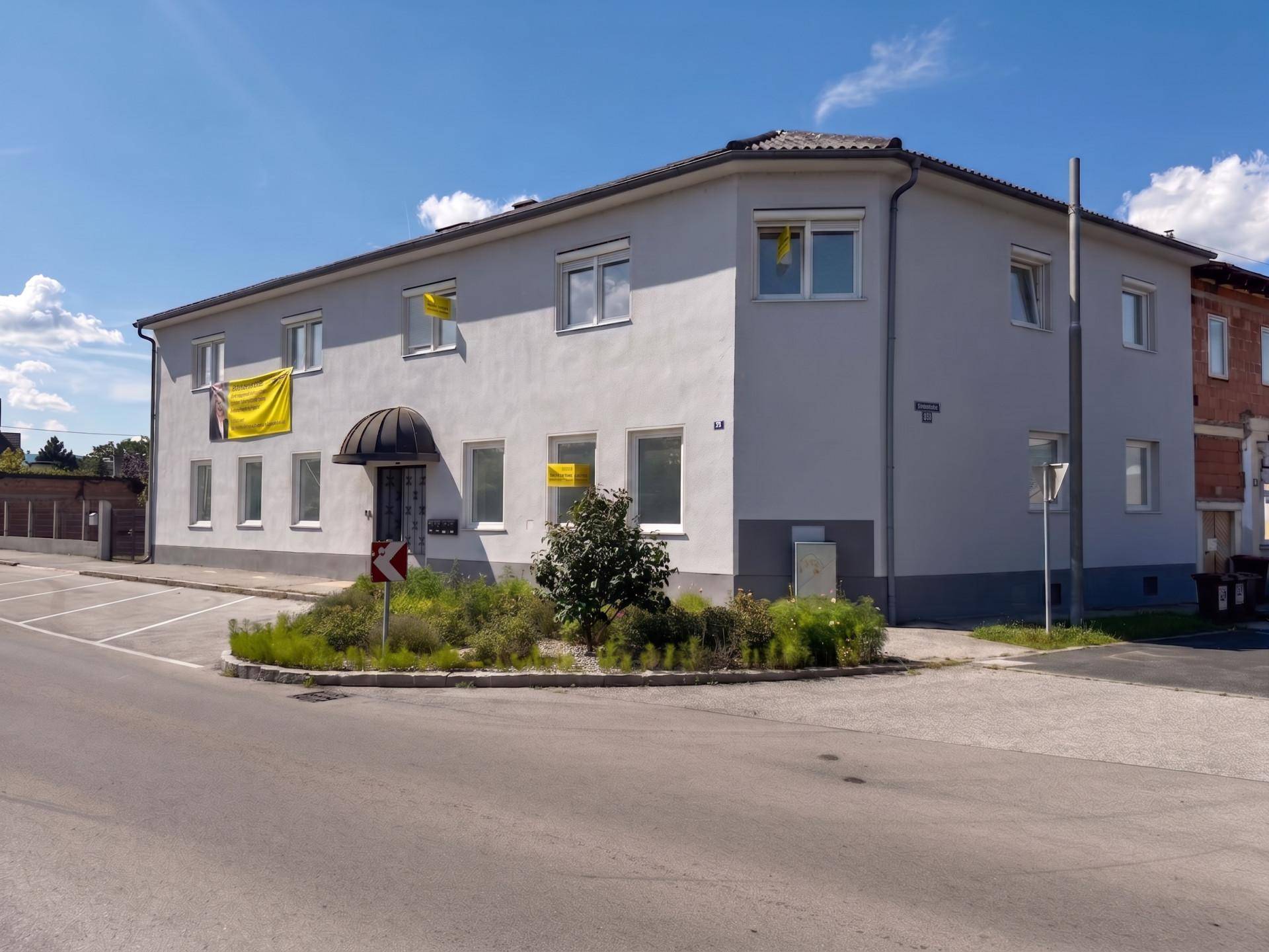 Außenansicht eines Mehrfamilienhauses mit heller Fassade und Parkplätzen davor.