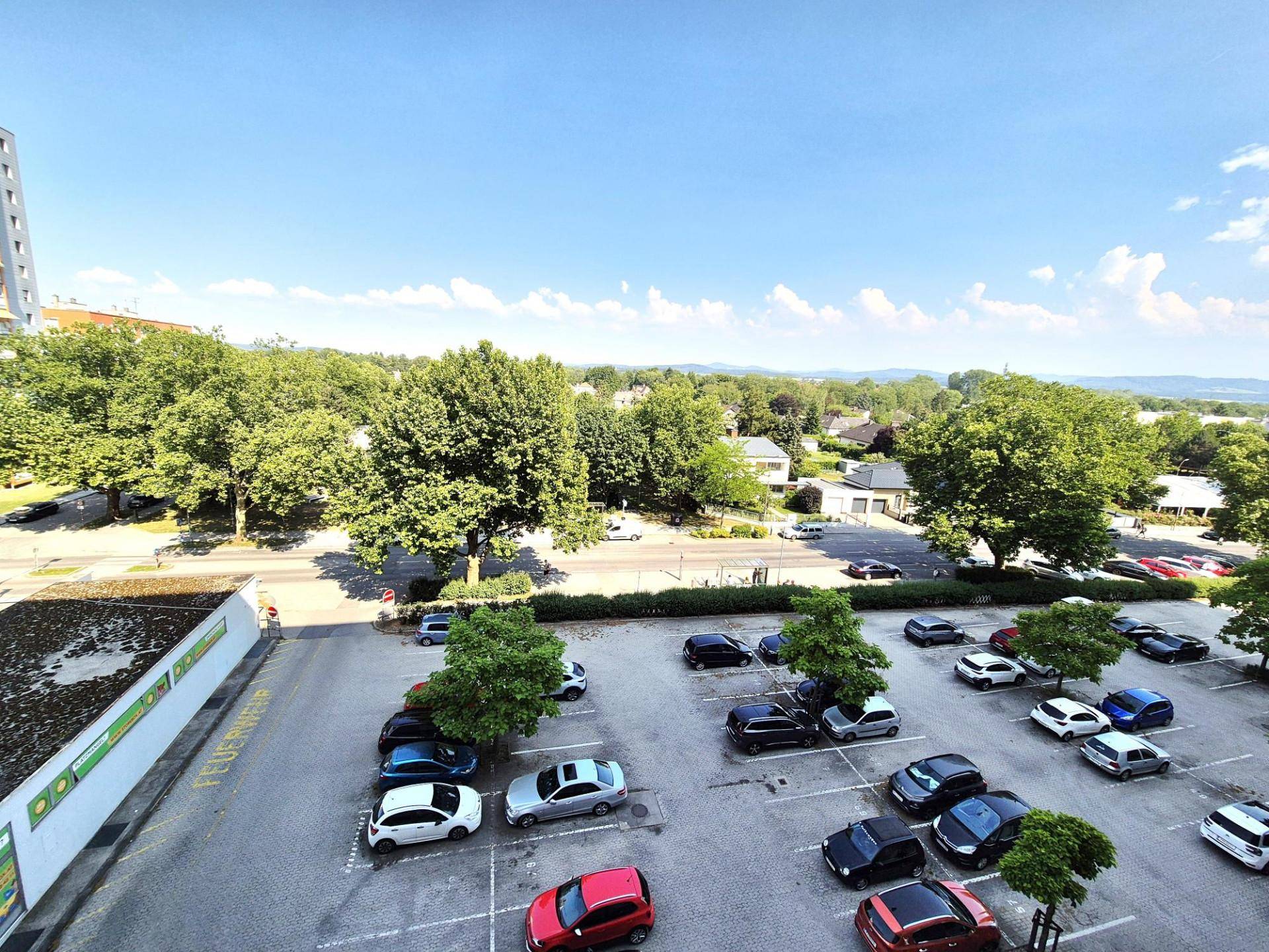 Großer Parkplatz mit vielen Autos und umliegenden Bäumen unter blauem Himmel.