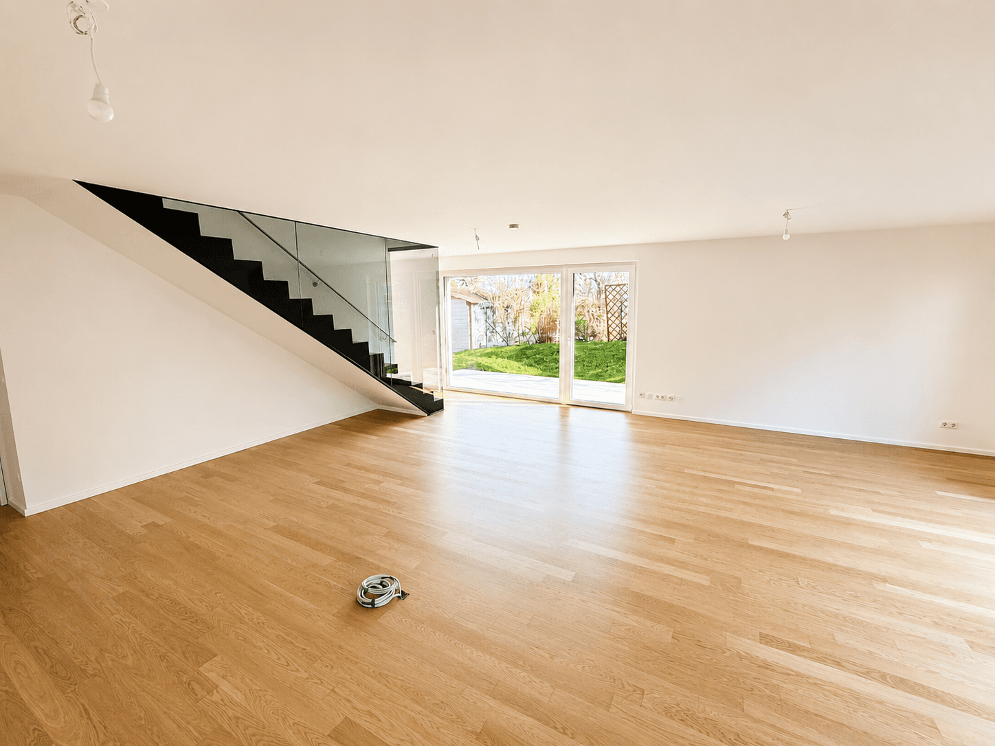 Geräumiges Wohnzimmer mit hellem Holzparkett, moderner Treppe und direktem Zugang zum grünen Außenbereich.
