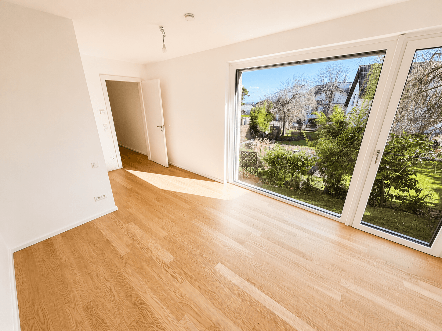 Geräumiger Raum mit hellem Holzboden und einer großen, schrägen Fensterfront mit Blick ins Grüne.