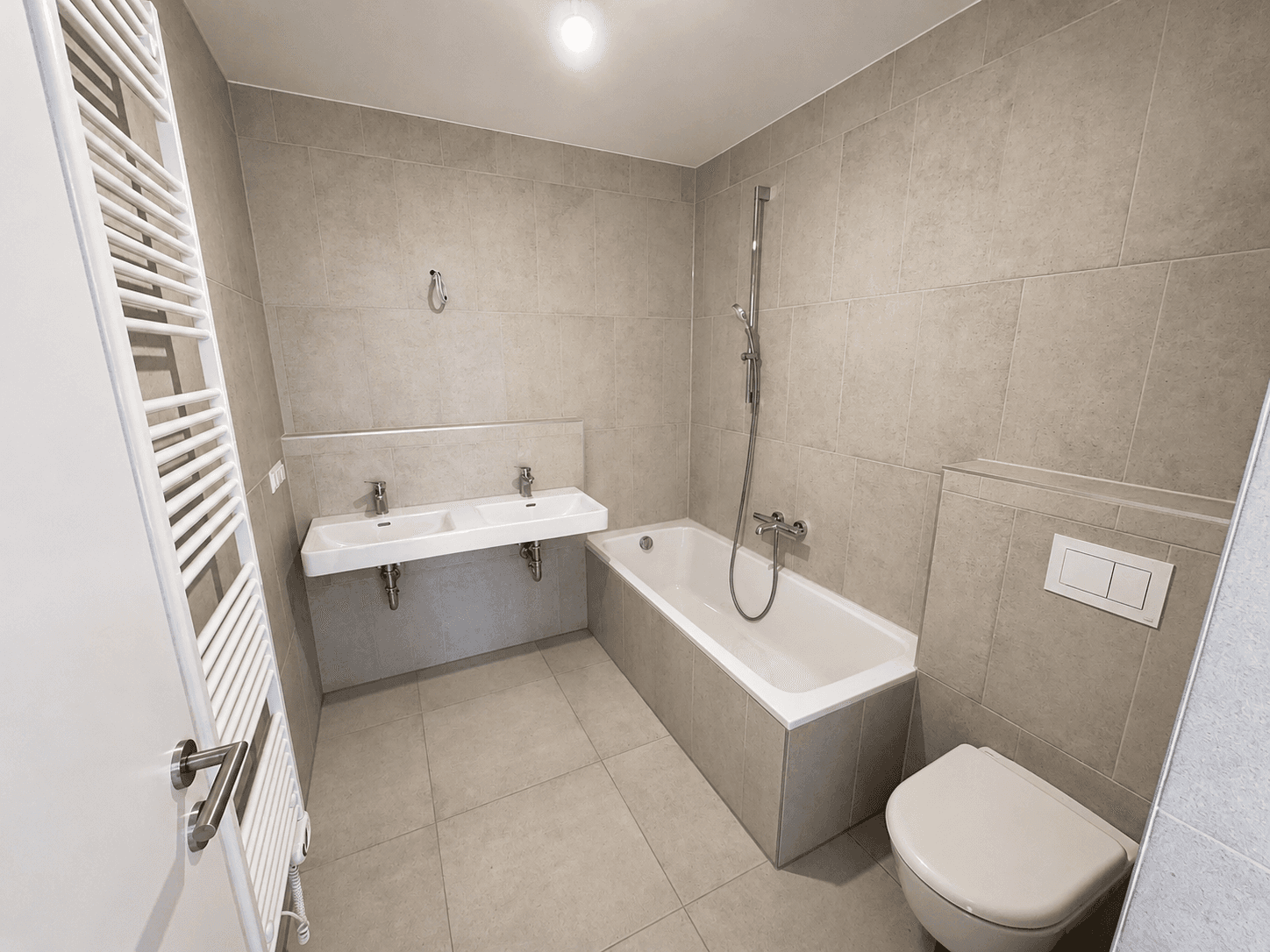 Modernes Badezimmer mit grauen Fliesen, Badewanne, Doppelwaschbecken und Wand-WC.