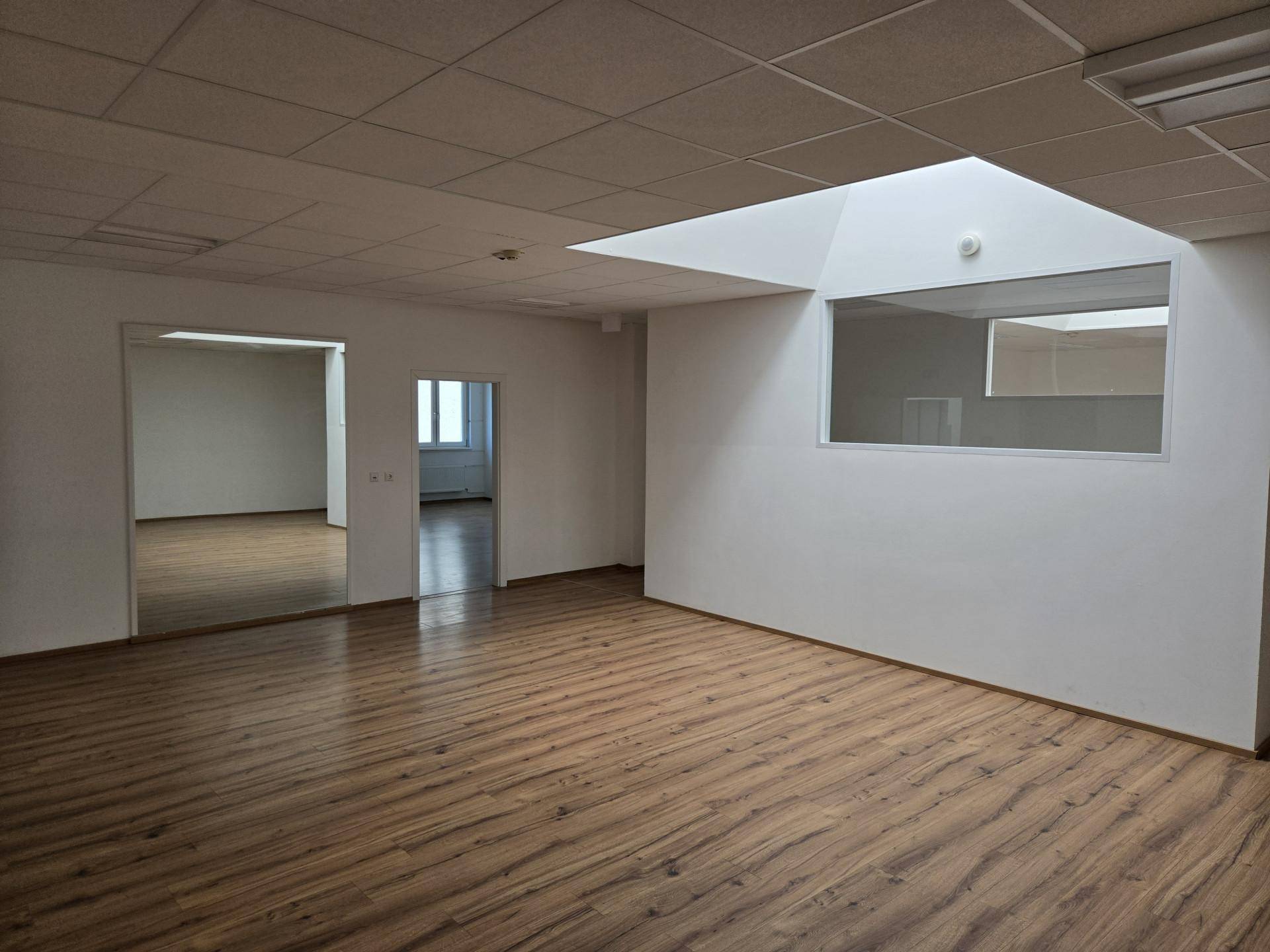 Großer Büroraum mit Laminatboden, abgehängter Decke und internem Fenster, angrenzend an weitere Räume.