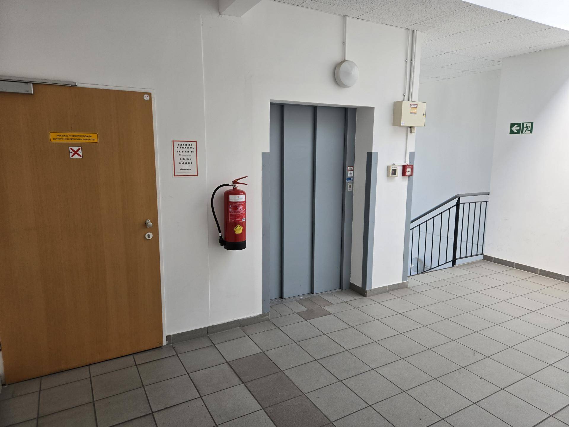 Flur mit Aufzug, Feuerlöscher, Holztür und Treppenhauszugang, ausgestattet mit Fliesenboden.
