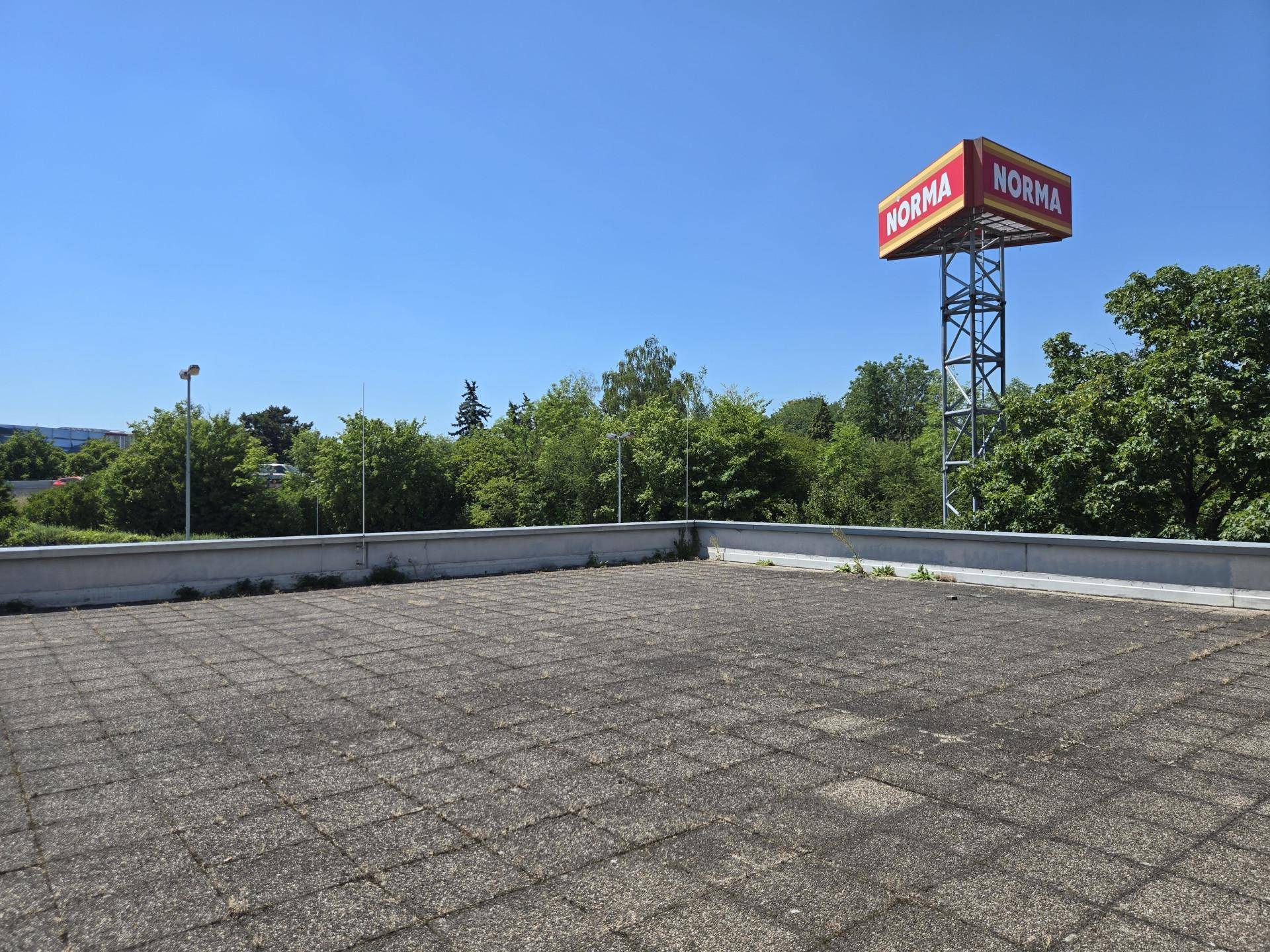 Weitläufige Dachfläche mit Betonplatten und Blick auf Bäume und ein Norma-Schild.