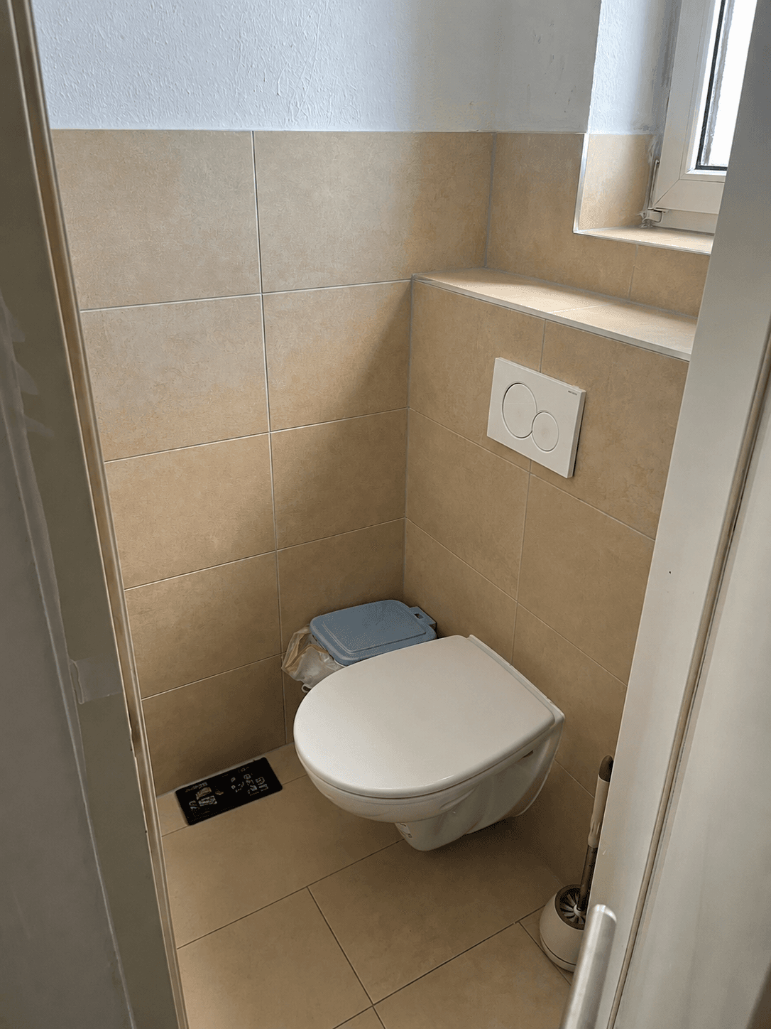 Separates WC mit wandhängender Toilette, Fenster und beigefarbenen Fliesen.