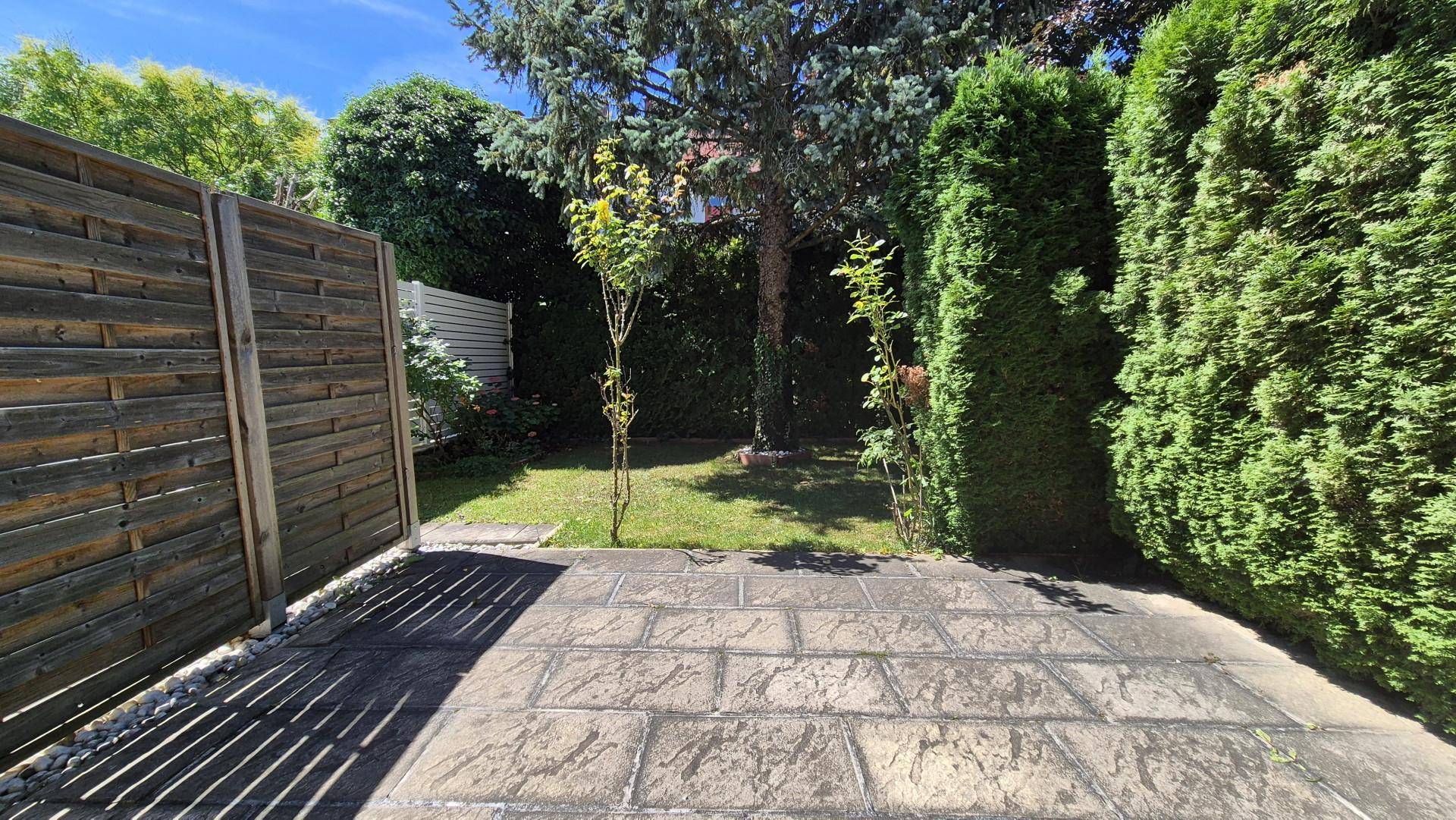 Sonniger Terrassenbereich mit Steinplatten und umgeben von grünen Hecken und Zäunen.