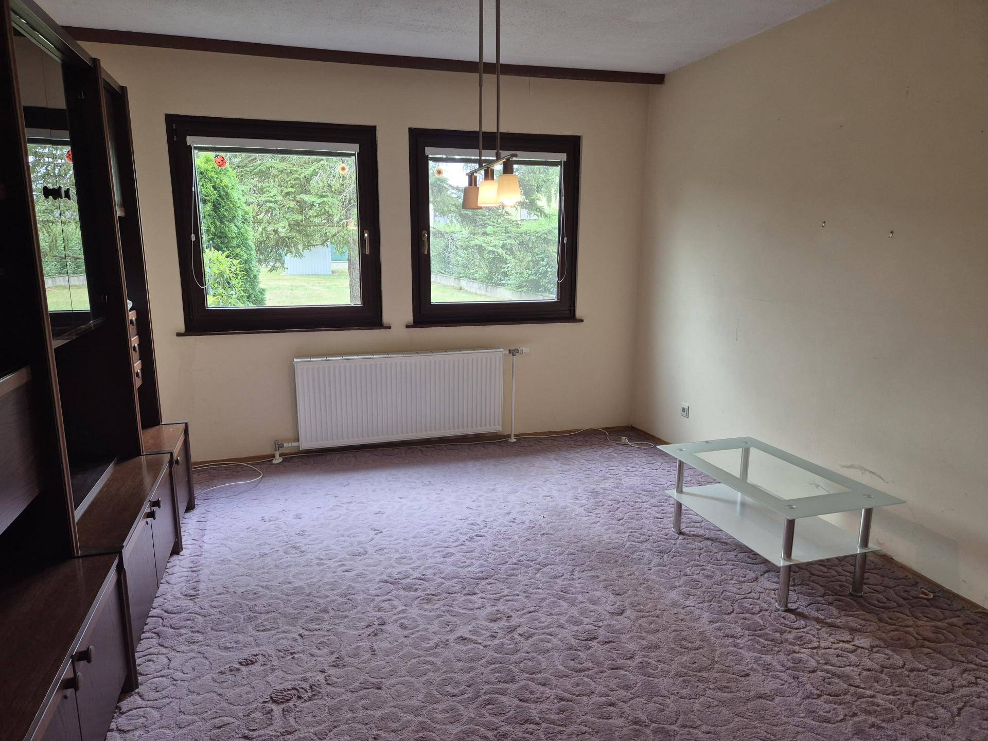 Geräumiges Wohnzimmer mit Teppichboden, großen Fenstern und Blick ins Grüne.