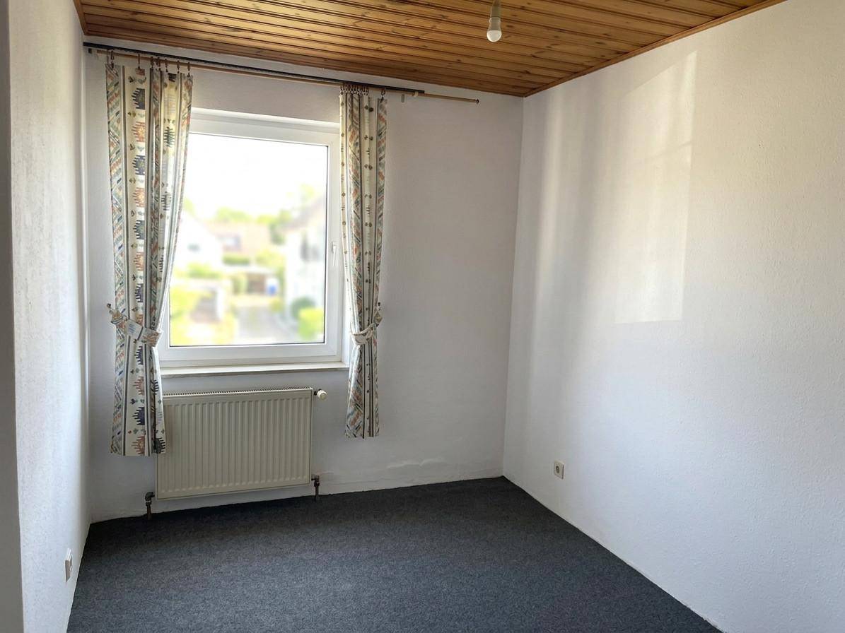 Leeres Zimmer mit dunklem Teppichboden, weißen Wänden und einem Fenster mit Blick nach draußen.