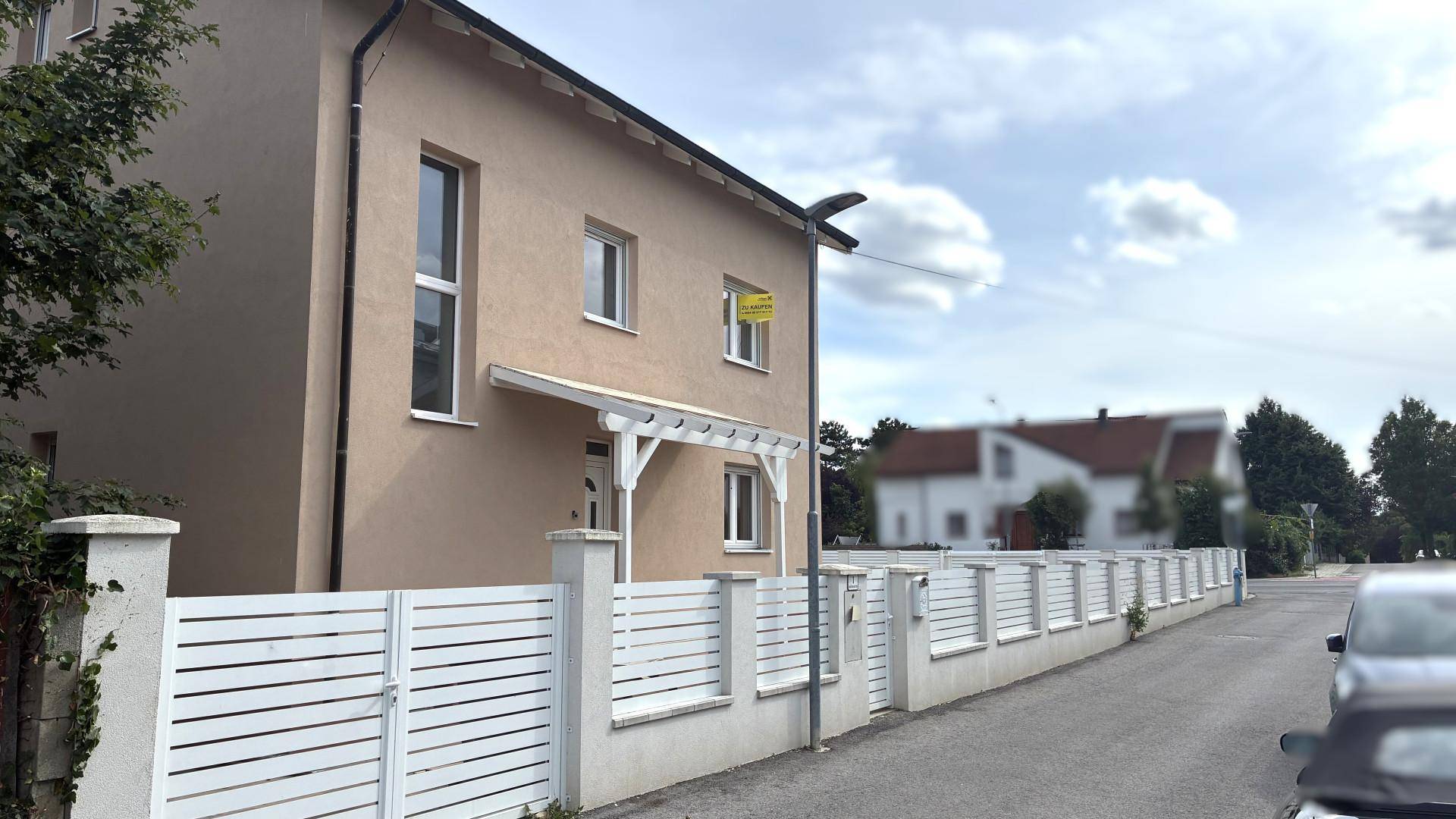 Modernes Haus mit heller Fassade, weißem Zaun und kleiner Pergola am Eingangsbereich.