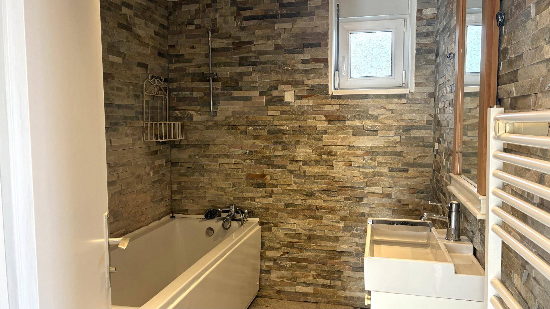 Modernes Badezimmer mit Steinoptik-Fliesen, Badewanne, Waschbecken und beheiztem Handtuchhalter.