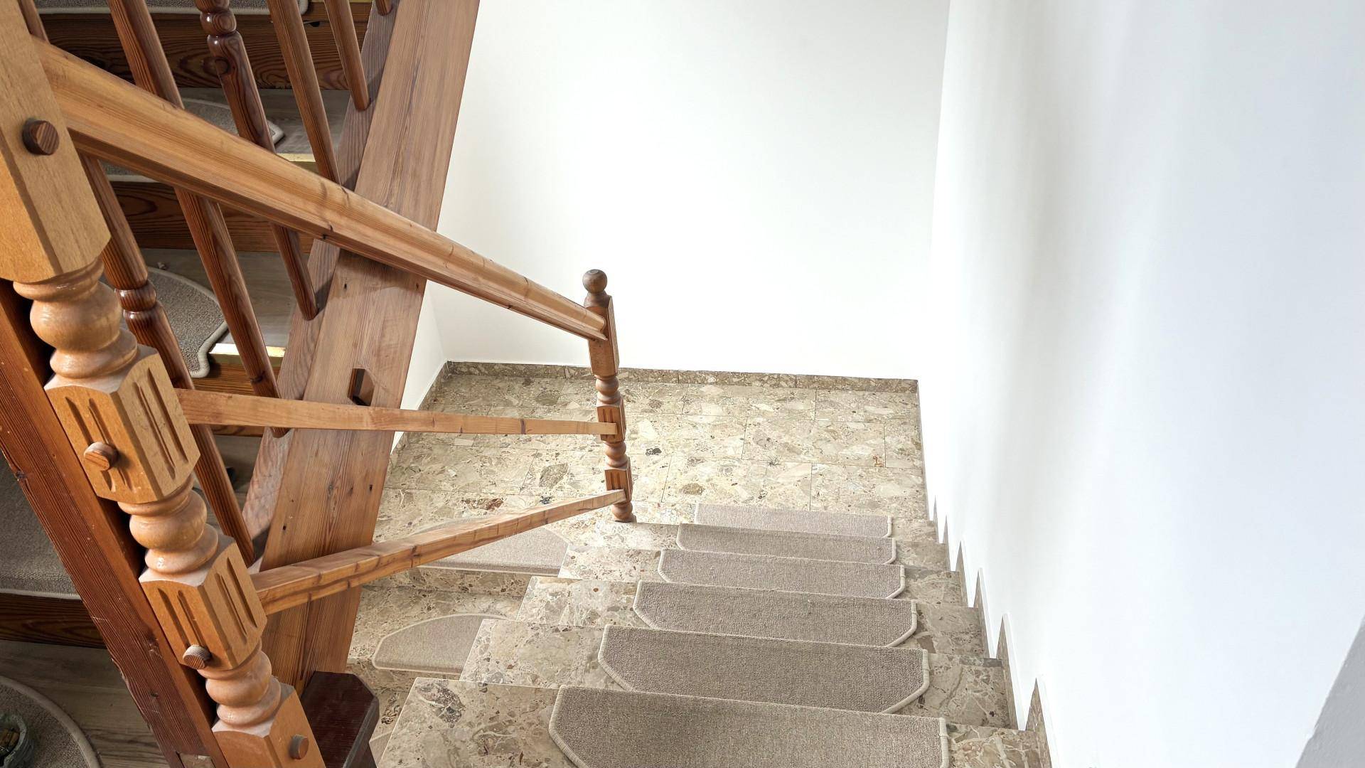 Blick von oben auf eine Treppe mit Holzgeländer und Teppichläufern auf Steinboden.