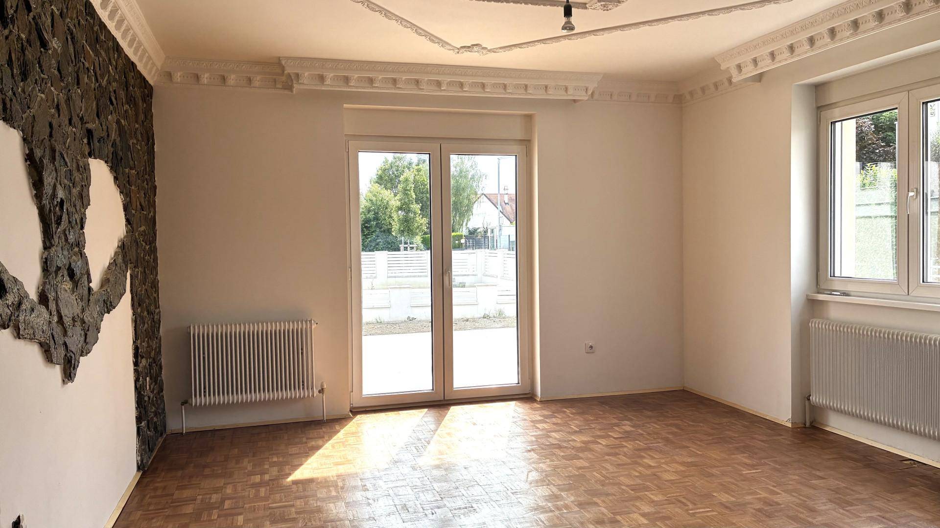 Geräumiger Raum mit Parkettboden, Stuckdecke, Heizkörper und großen Türen zum Außenbereich.