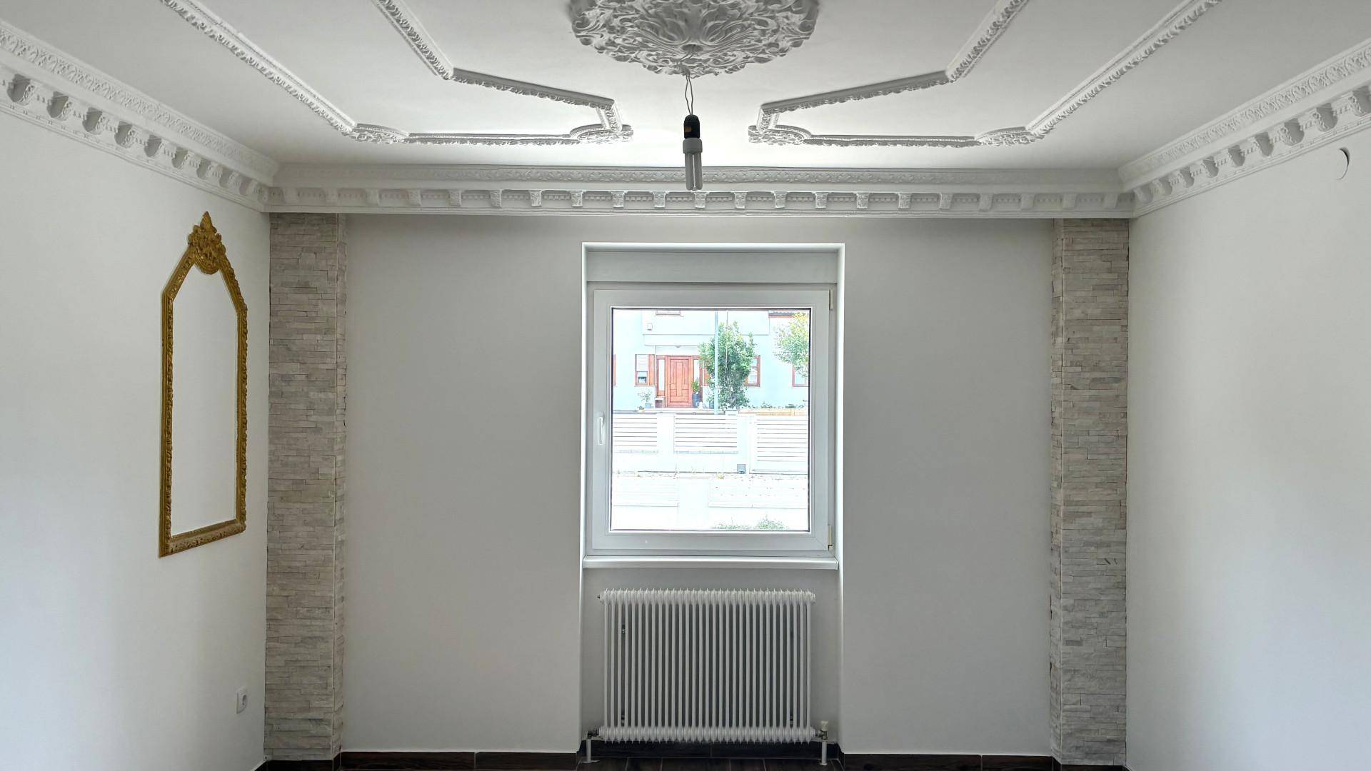 Heller Raum mit weiß gestrichenen Wänden, Fenster, Heizkörper und dekorativer Stuckdecke.