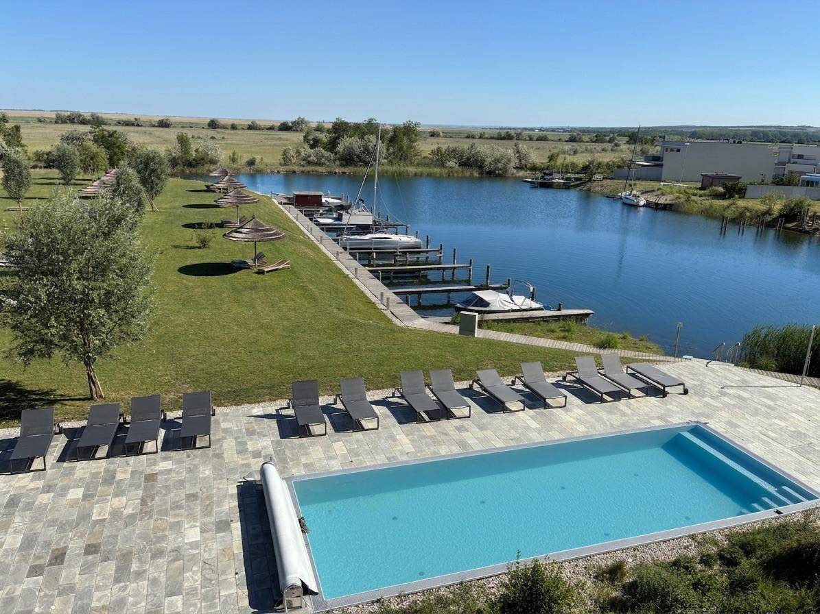 Panoramablick auf den Poolbereich mit Liegestühlen, angrenzendem See und Bootsanlegestellen.