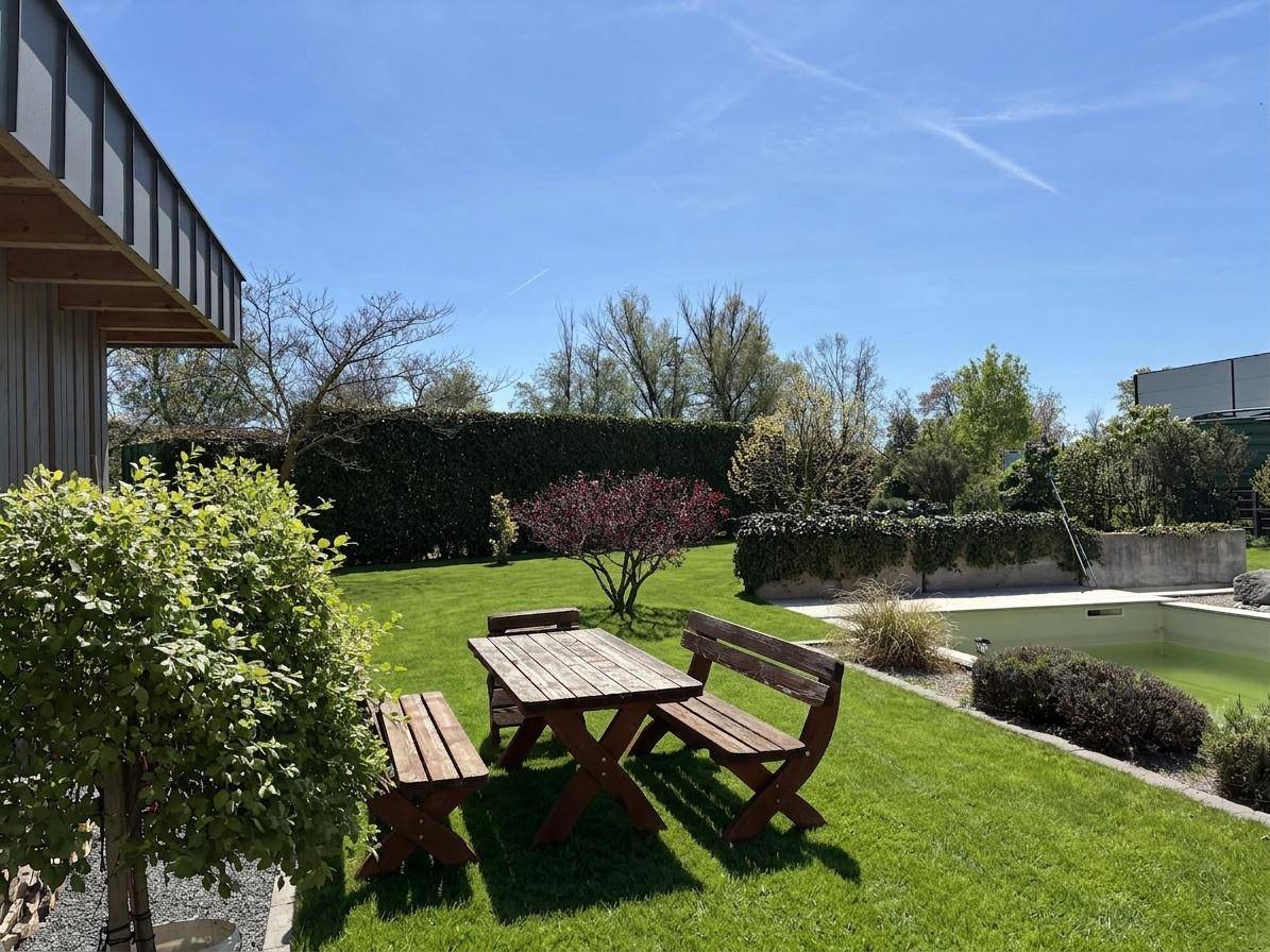 Sonnige Gartenecke mit rustikalen Holzmöbeln und einem kleinen Poolbereich im Hintergrund, umgeben von Grün.