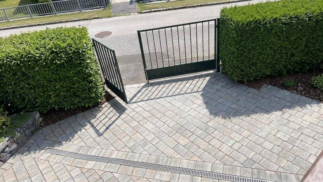 Gepflasterter Eingangsbereich mit offenem Metalltor und hohen, gepflegten Hecken zur Straßenseite.