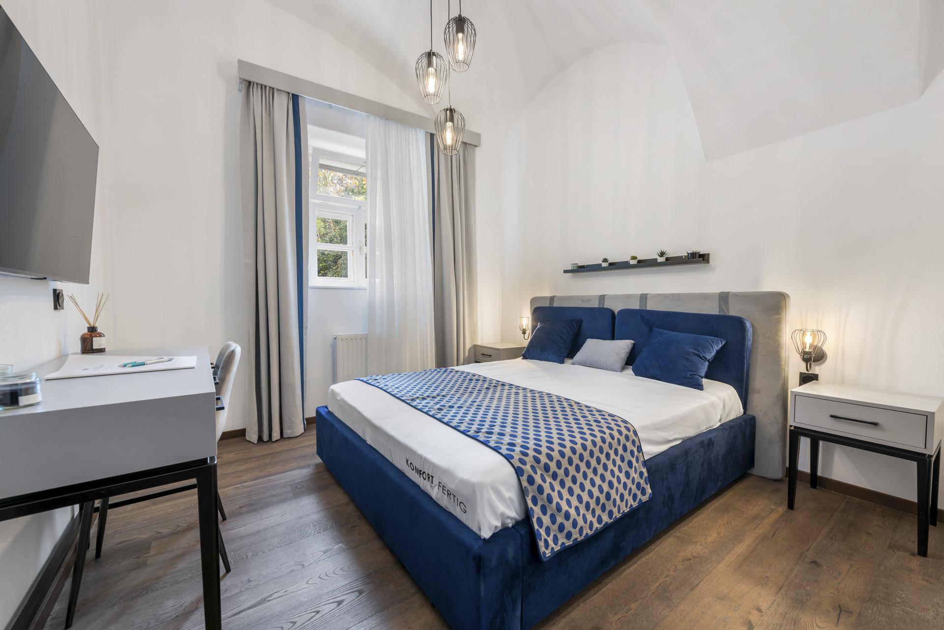 Helles Schlafzimmer mit blauem Doppelbett, Schreibtisch und Nachttischen unter einem Gewölbedach.