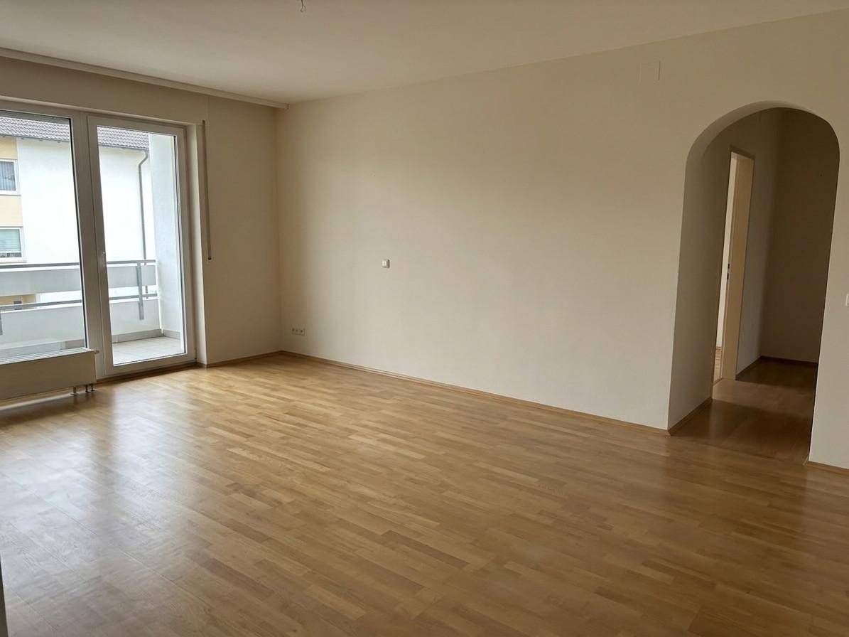 Geräumiges Wohnzimmer mit Parkettboden, Balkonzugang und offenem Durchgang zu einem weiteren Raum.