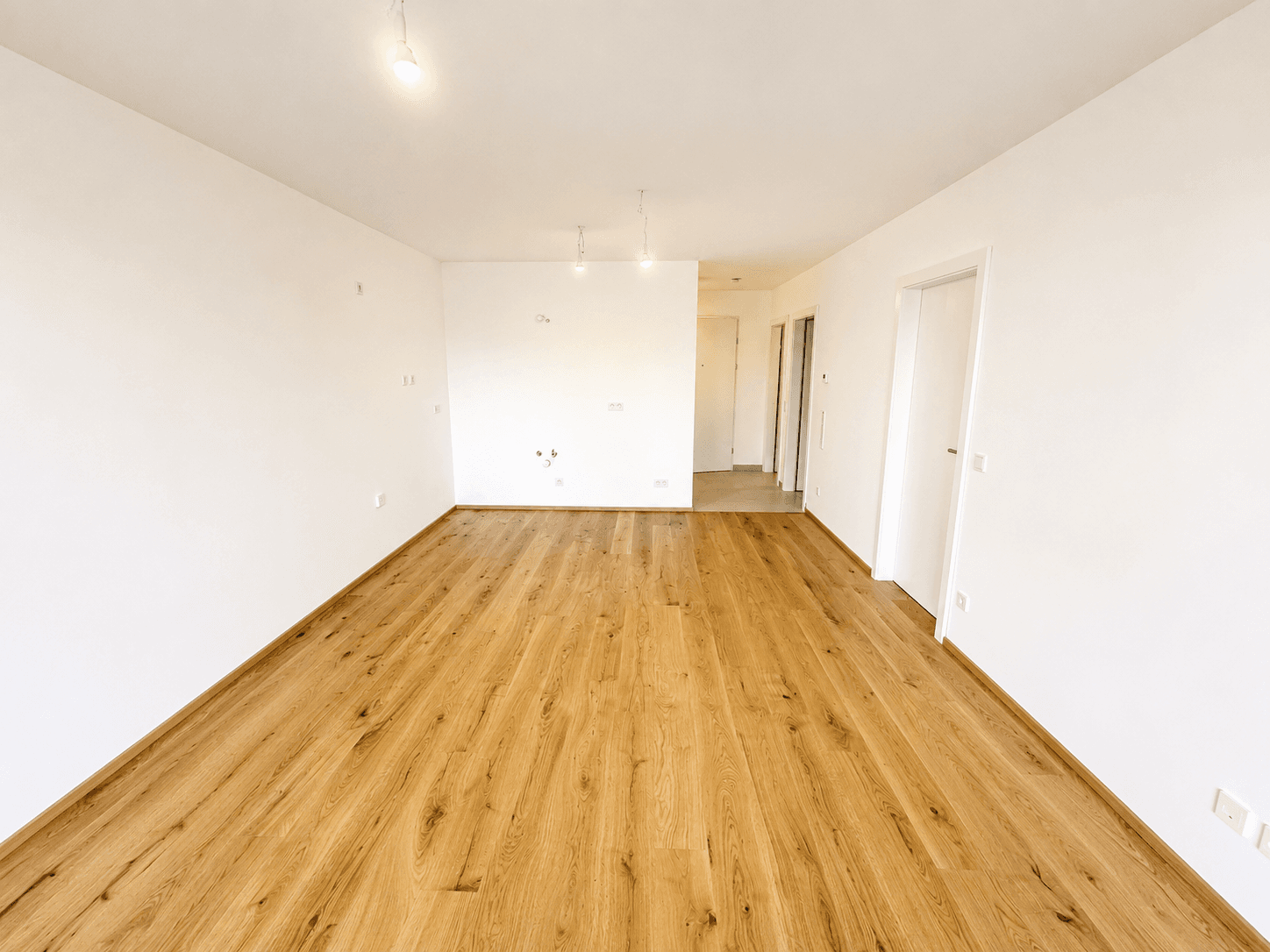 Leerer Raum mit Parkettboden und Anschlüssen an der Wand, bereit für die Kücheninstallation.