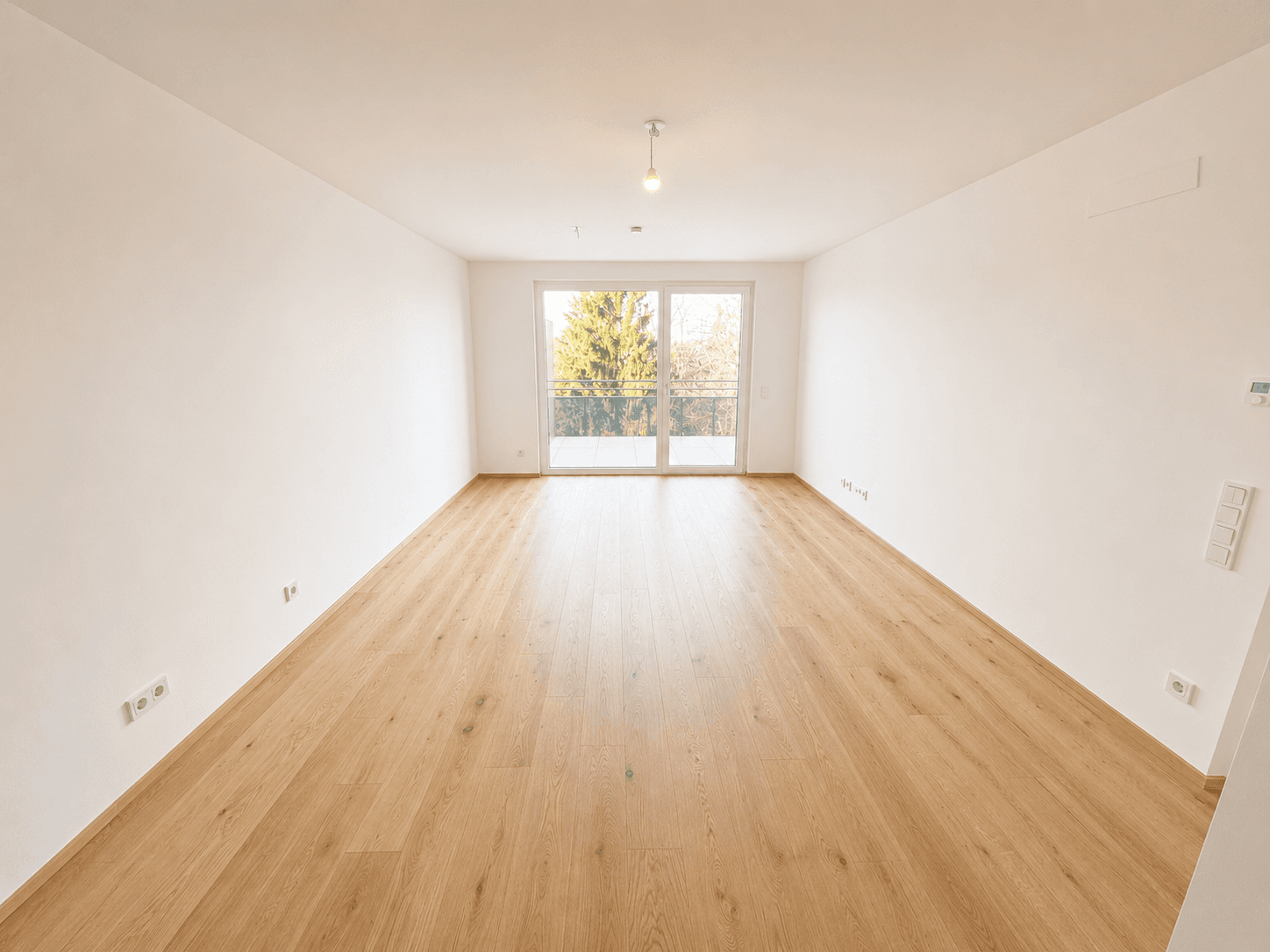 Heller, leerer Wohnraum mit Laminatboden und großen Glasfenstern, die Zugang zu einem Außenbereich bieten.