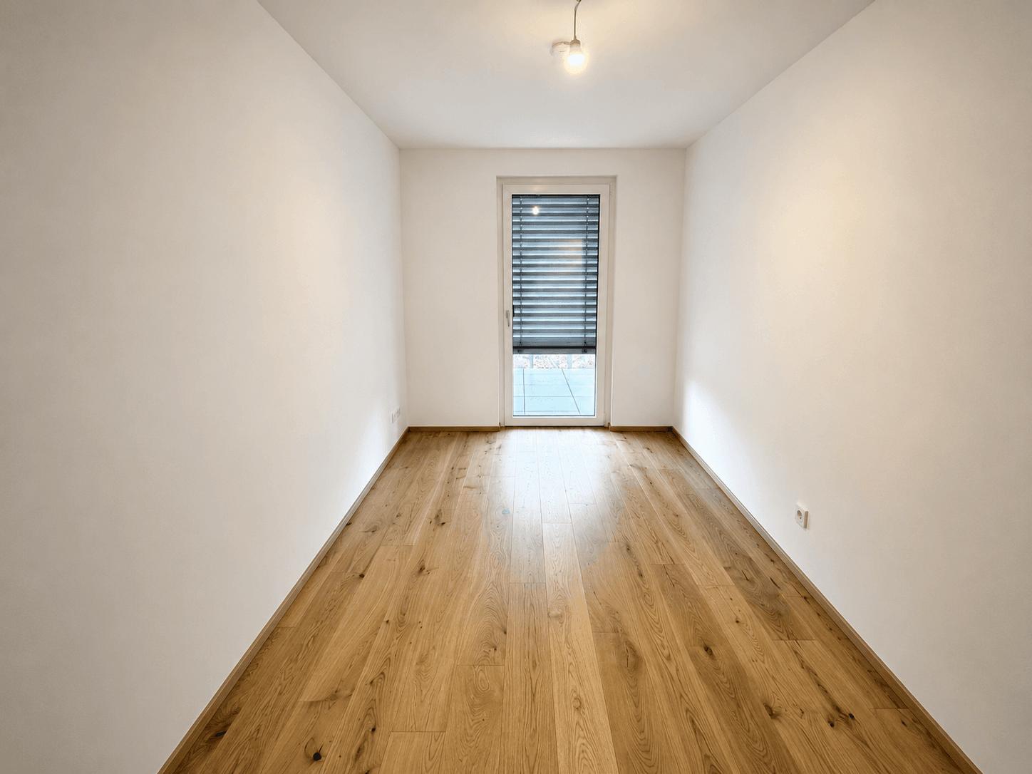 Leeres Zimmer mit Laminatboden, weißen Wänden und einer Tür mit Rollladen, die nach draußen führt.