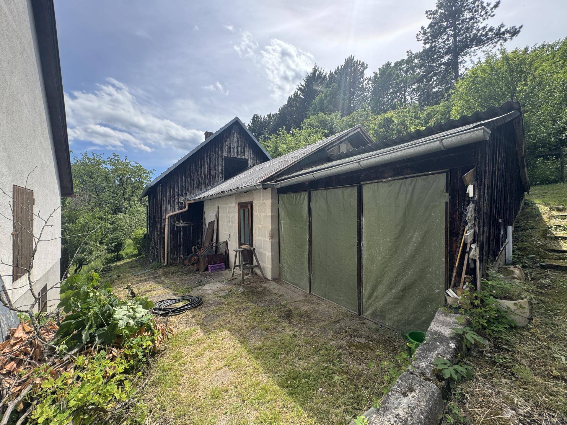 Anbau mit Garagentoren und angrenzendem Holzgebäude in ländlicher Umgebung, umgeben von Grün.