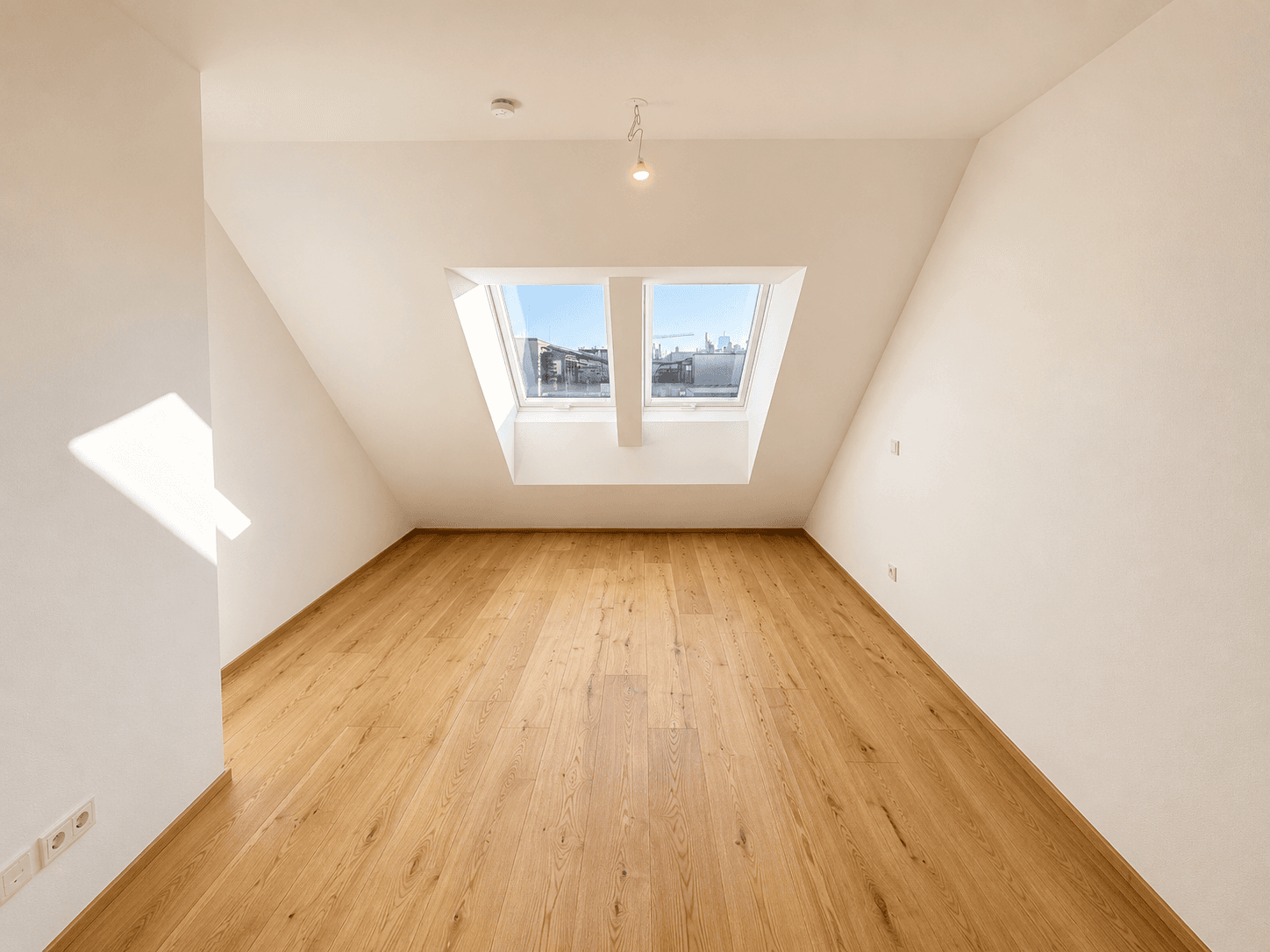 Lichtdurchflutetes Dachzimmer mit Holzboden, schrägen Wänden und zwei großen Dachfenstern.